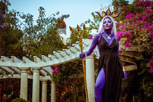 A model in elaborate fantasy costume poses outdoors in a garden backdrop.