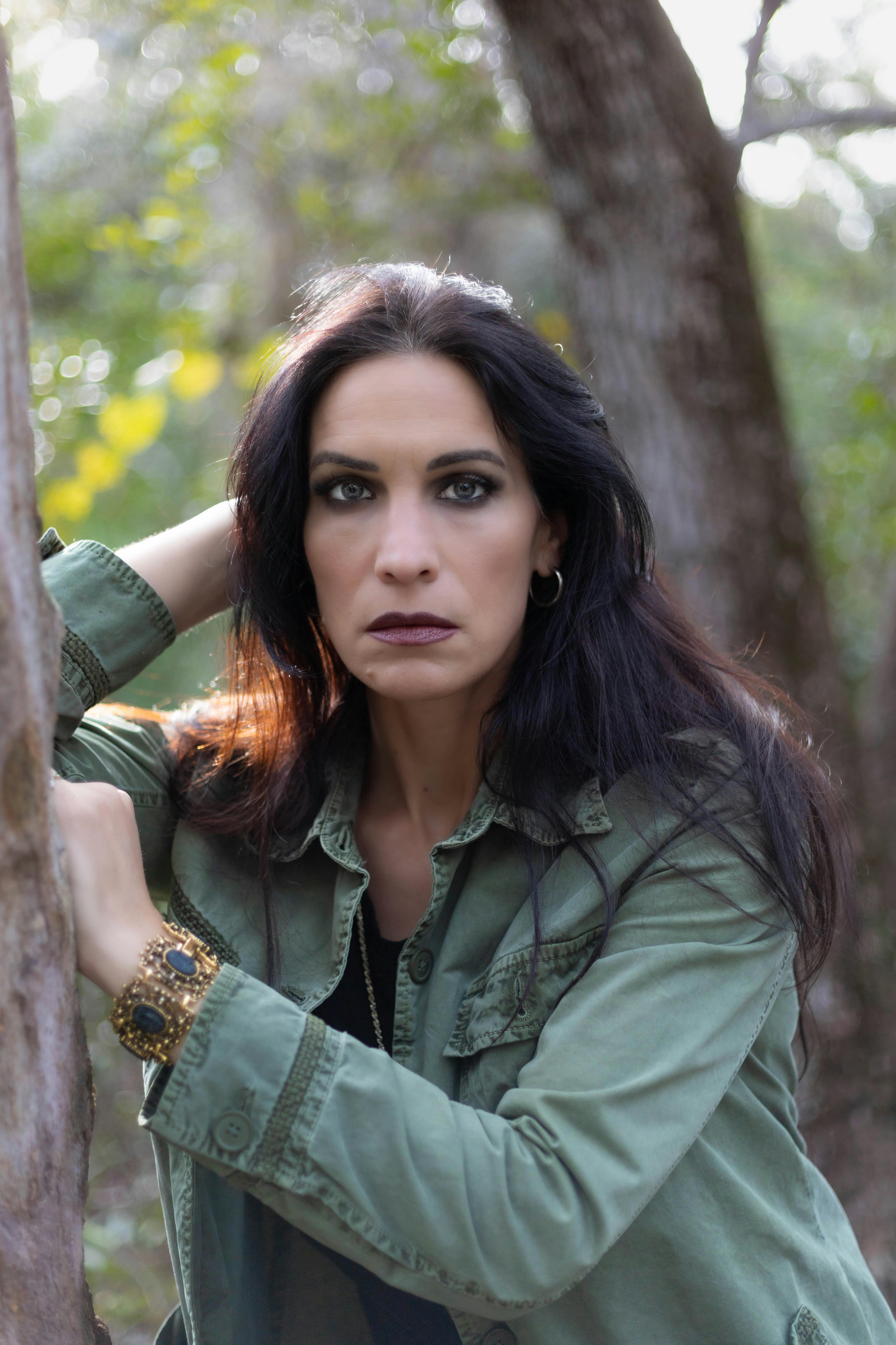 Elegant woman in a green jacket with dramatic makeup posing thoughtfully in a forest setting.