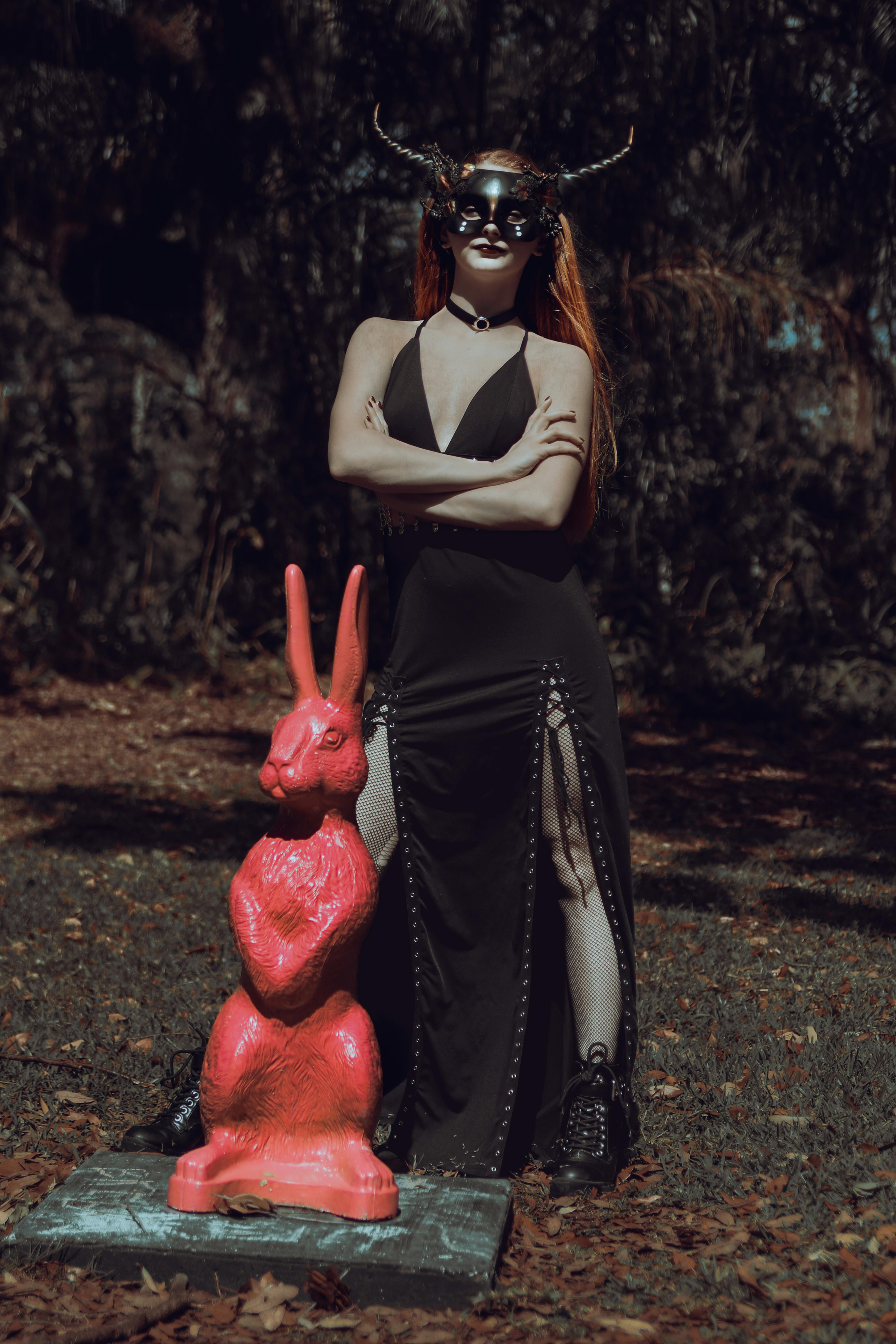 A woman wears a horned mask and black dress next to a red sculpture outdoors.