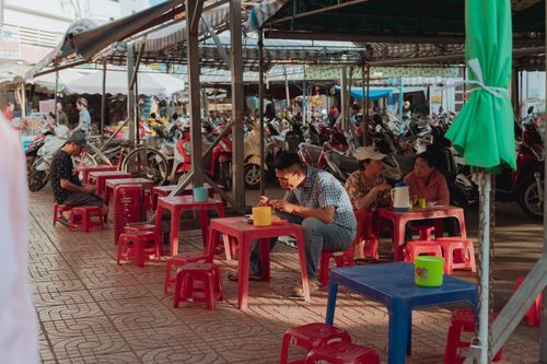 Free Casual outdoor dining in a Vietnamese market setting with vibrant plastic stools and people enjoying food. Stock Photo