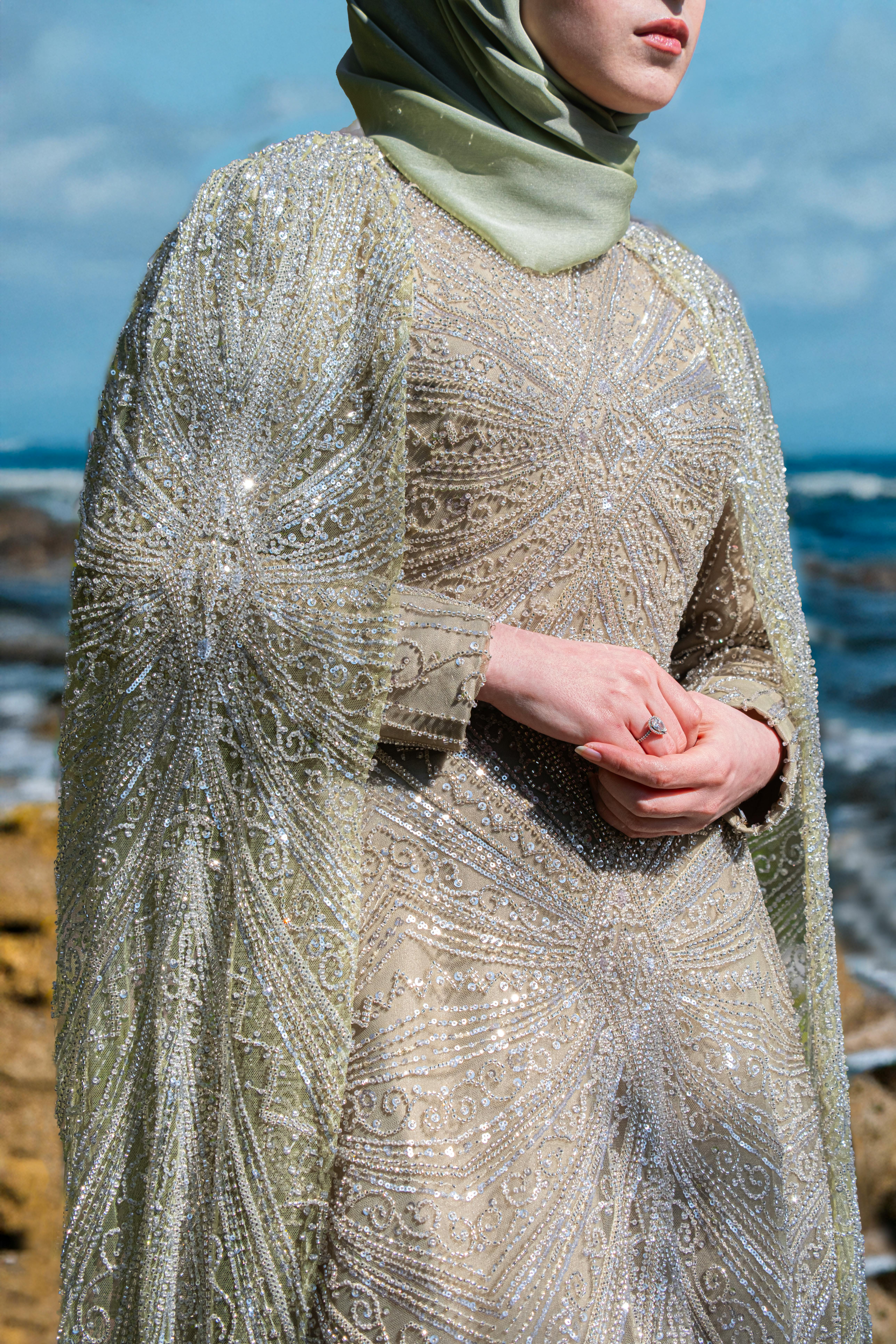 A woman in a glittering beaded dress stands elegantly by the ocean.