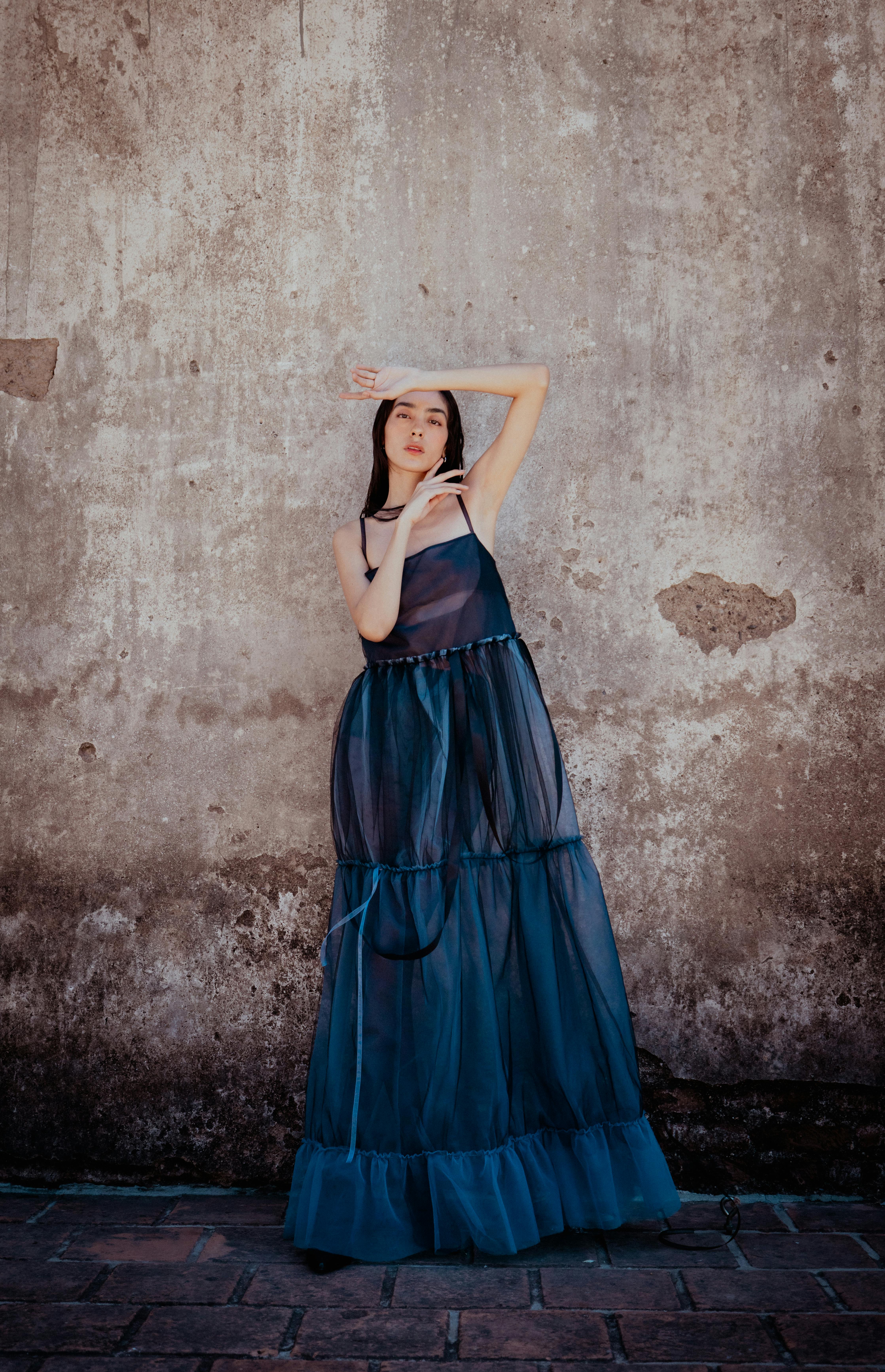 Elegant woman in a blue dress poses against a rustic wall in León, Mexico, showcasing beauty and fashion.