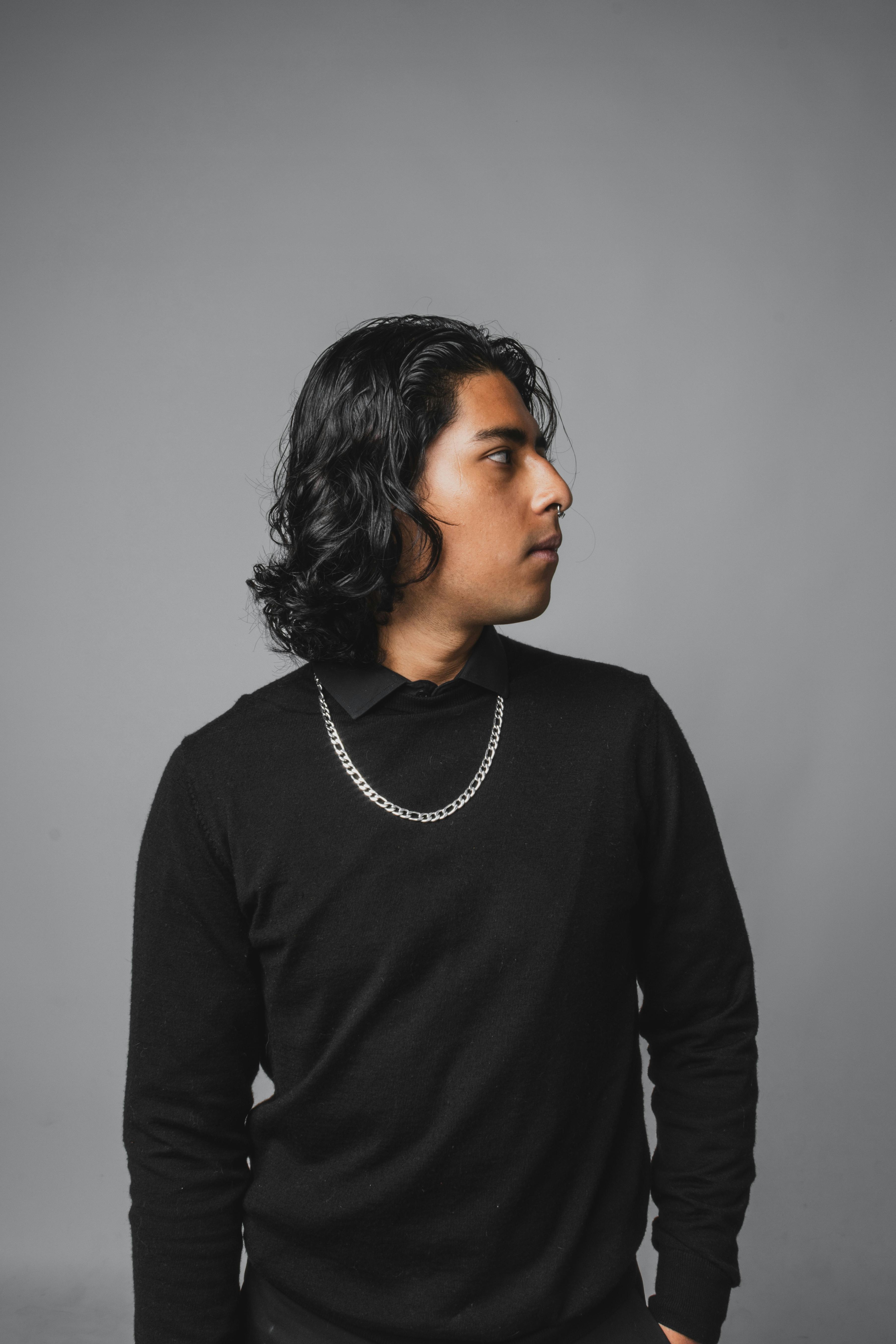 gratis Portret van een stijlvolle jongeman met lang zwart haar en een ketting, die zijdelings naar binnen kijkt. Stockfoto