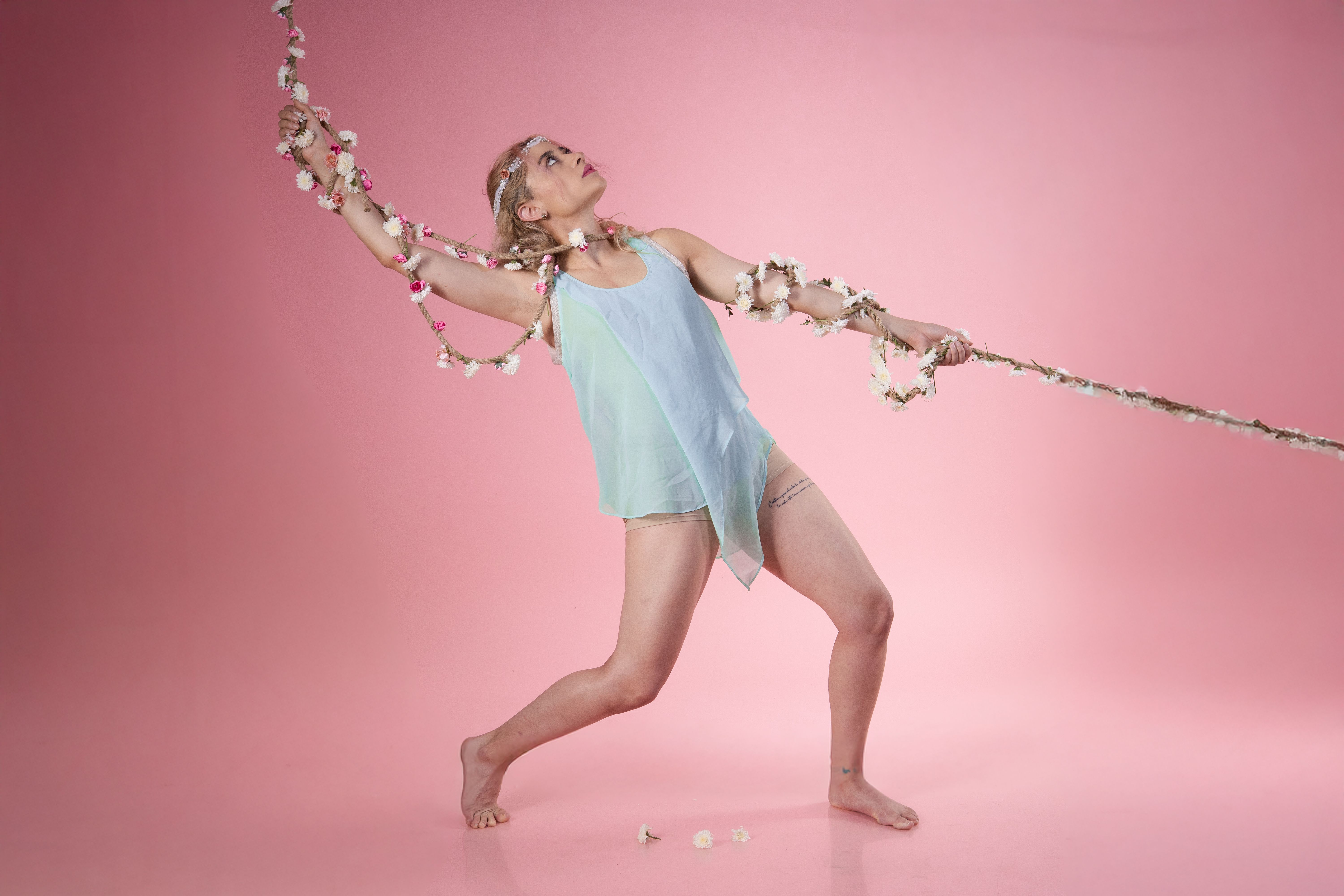 A blonde dancer poses gracefully with a flower-adorned rope against a pink background.