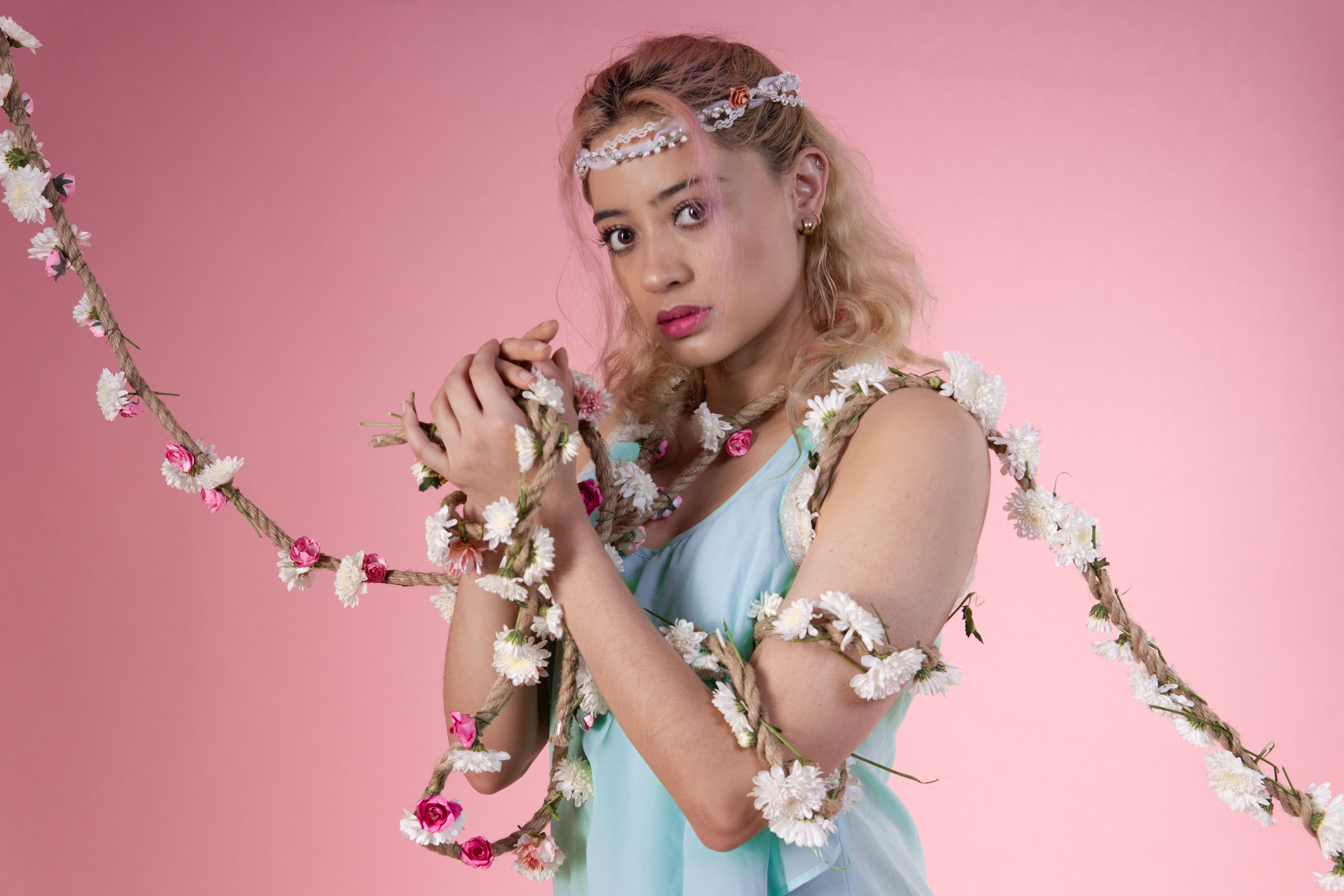 Blonde dancer poses with a floral garland against a pink background, exuding grace and elegance.