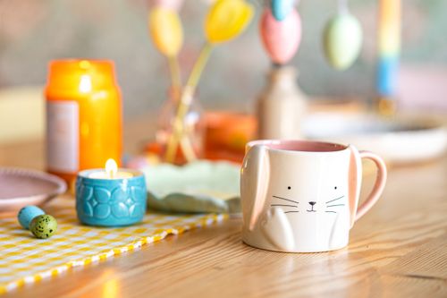 Free Easter-themed setup with a bunny mug, candle, and colorful decor on a wooden table. Stock Photo