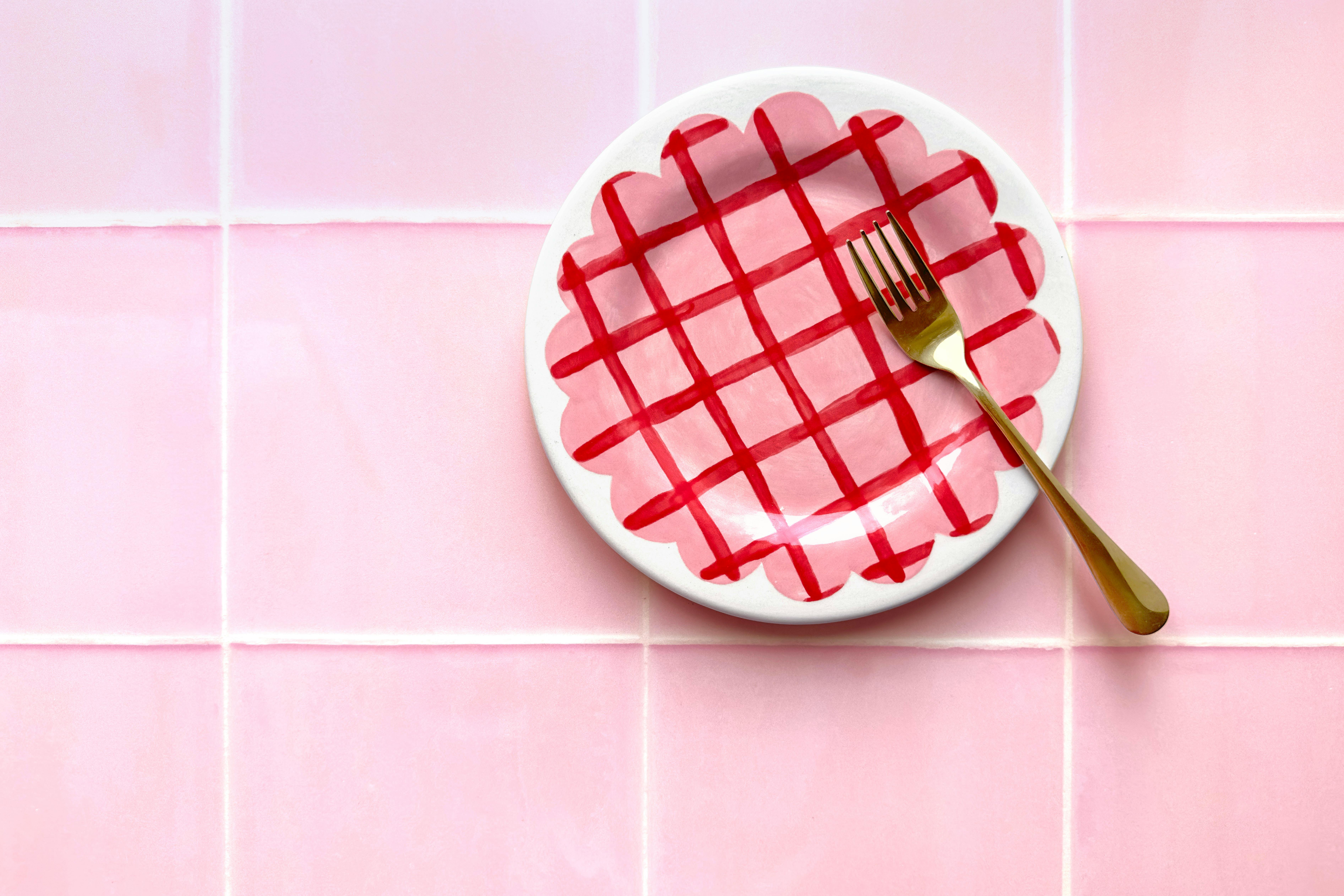 A pink ceramic plate with a red checkered pattern and a fork on a tiled surface.