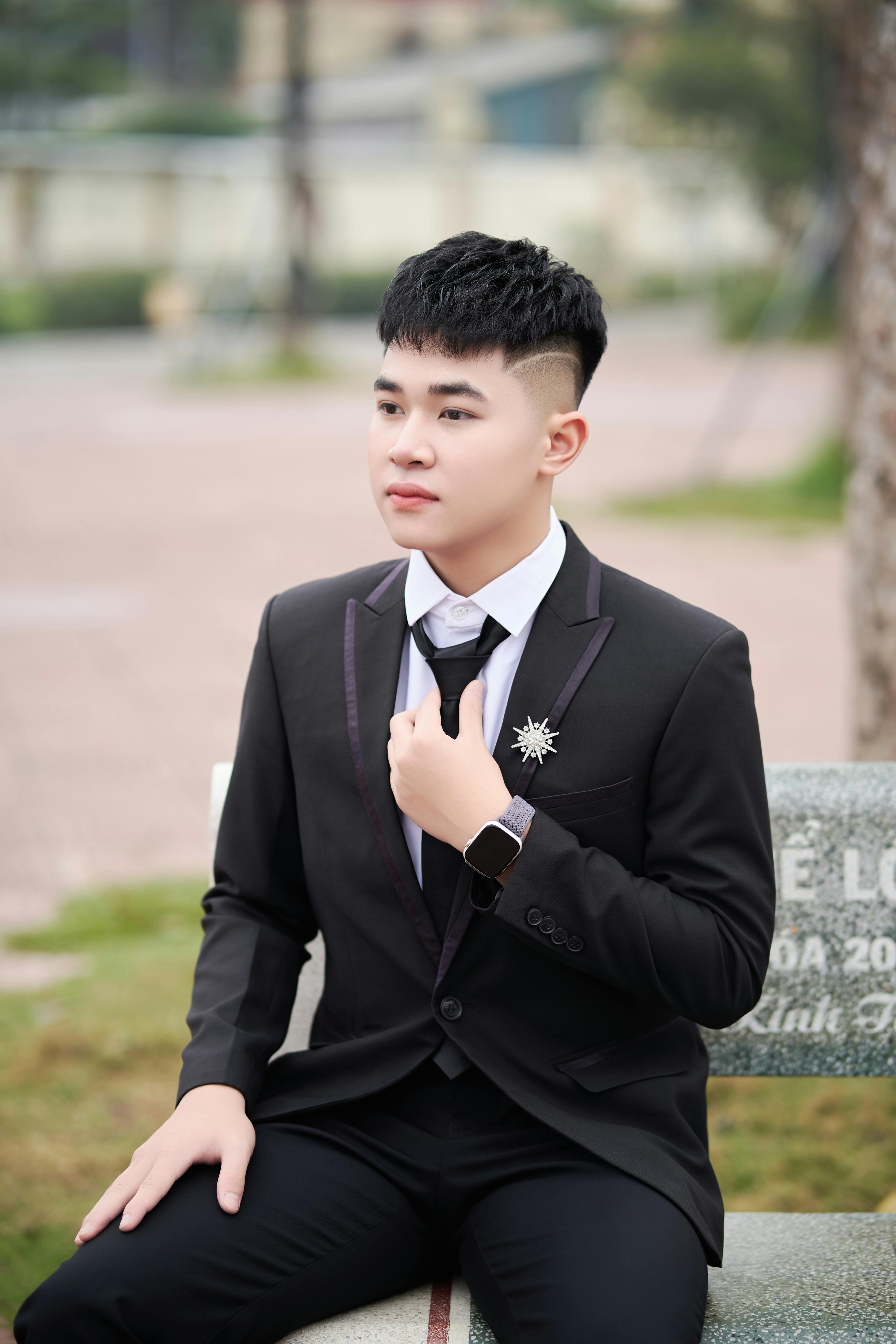 Stylish young man in a black suit posing outdoors on a bench, exuding confidence and elegance.