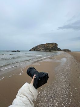 A serene beach scene with a person holding a camera, capturing a rocky island.