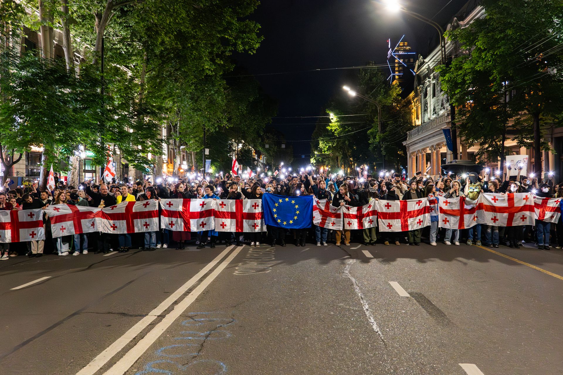 A large nighttime protest in Tbilisi with people holding Georgian and EU flags.