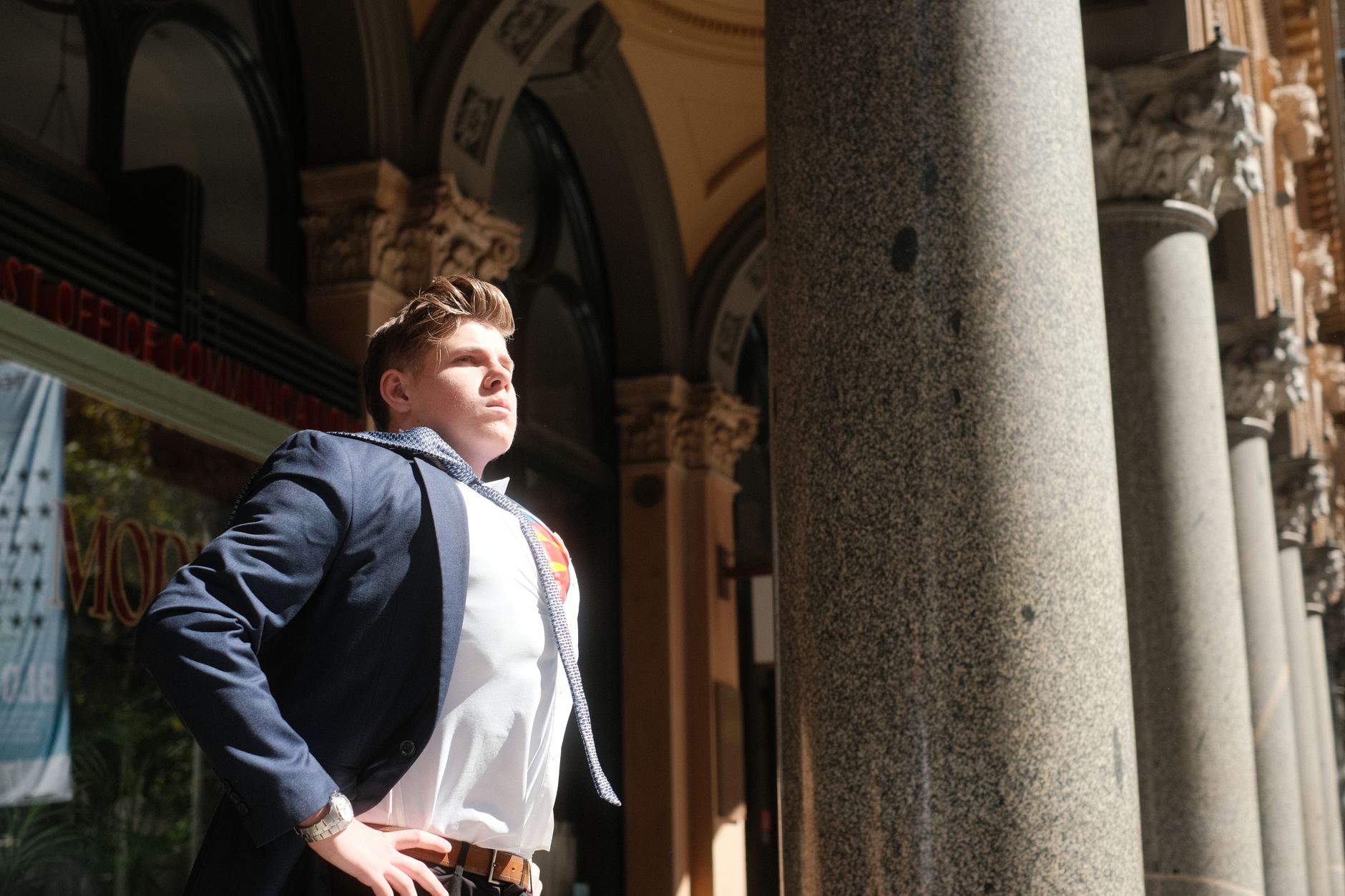 A determined man in a blue suit poses heroically outdoors under bright sunlight in Sydney, Australia.