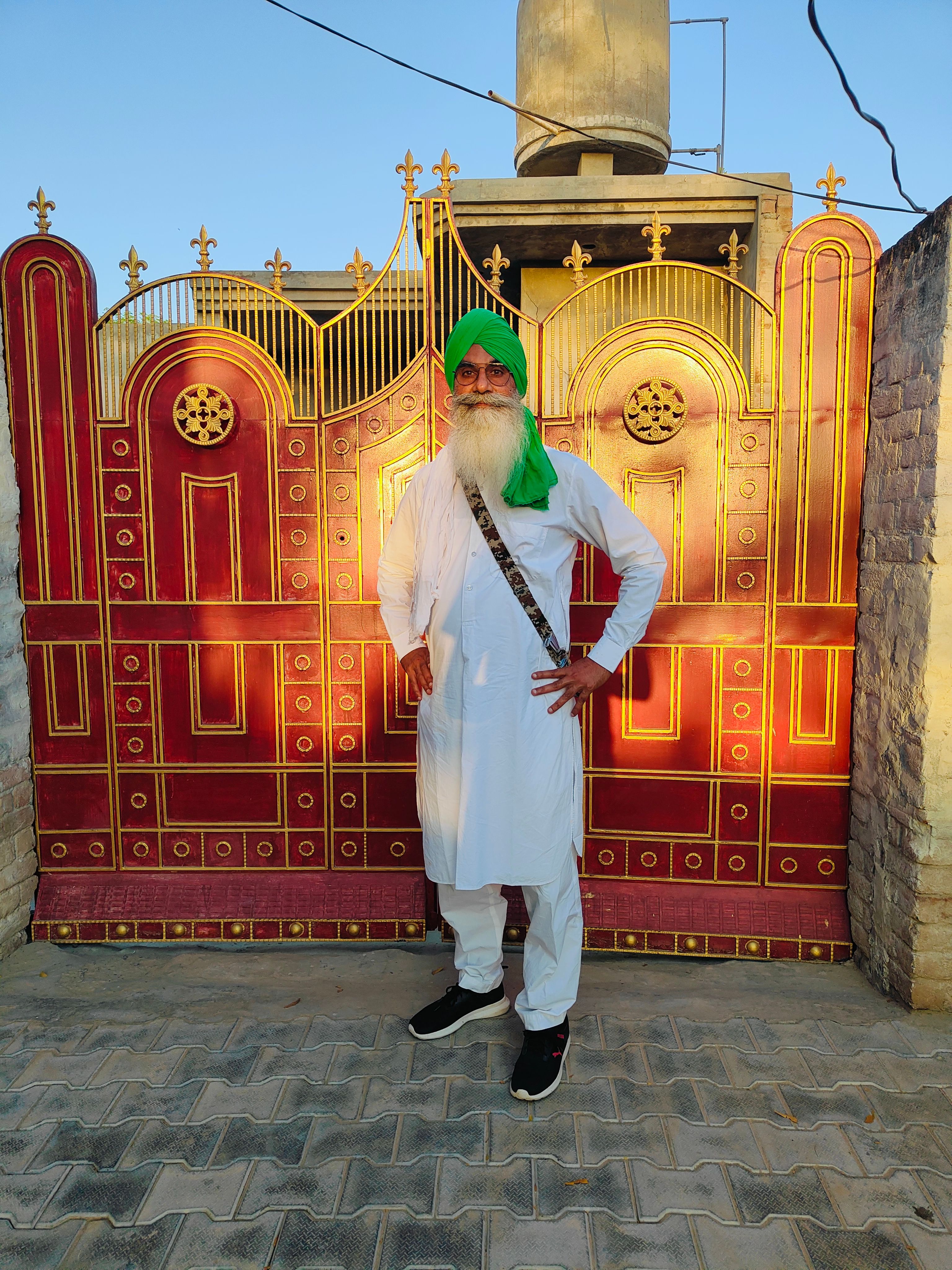 Elderly man in white attire and green turban standing by a decorative gate in daylight.