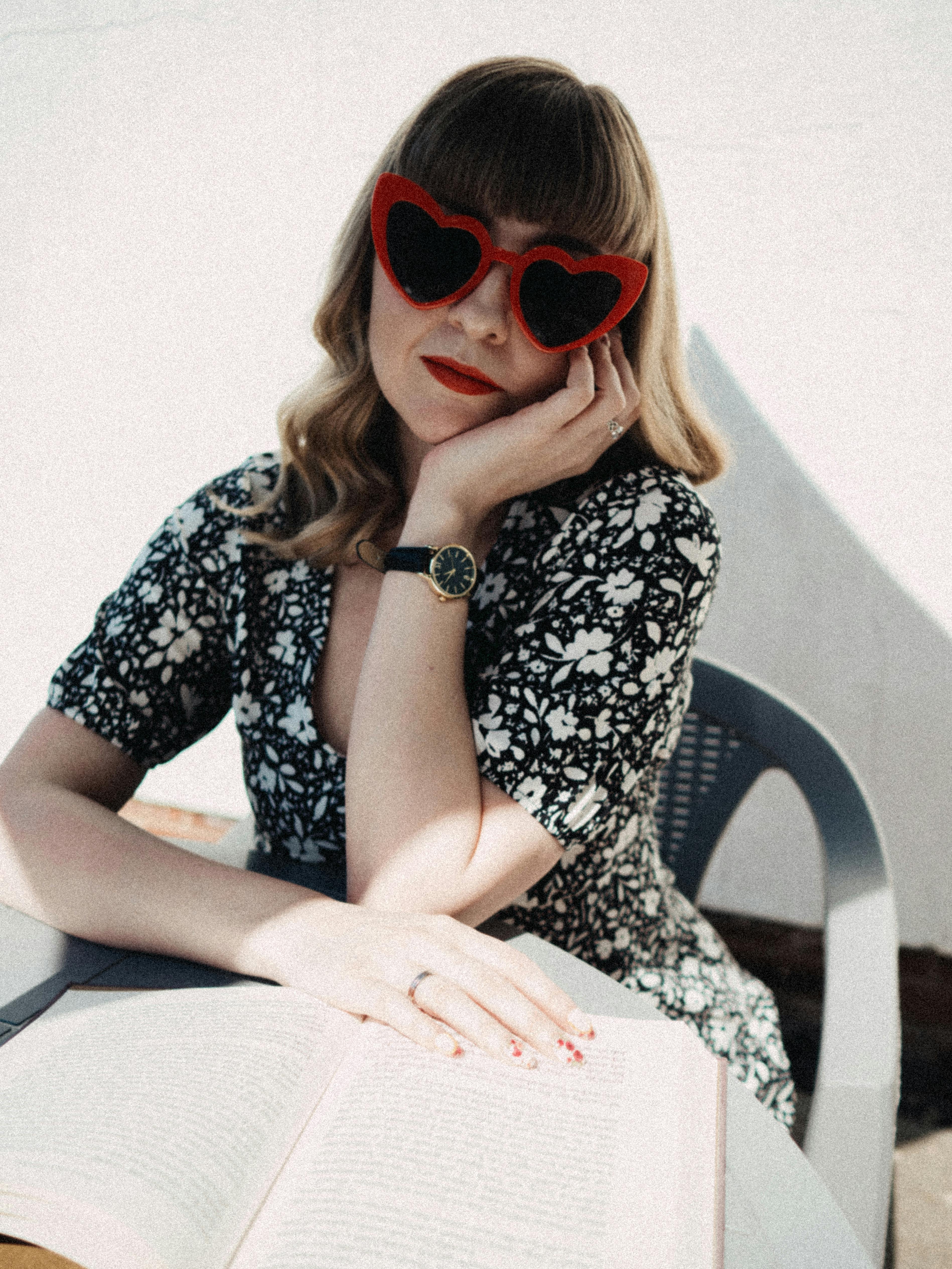 Trendy woman in floral dress with heart-shaped sunglasses reading a book outdoors.
