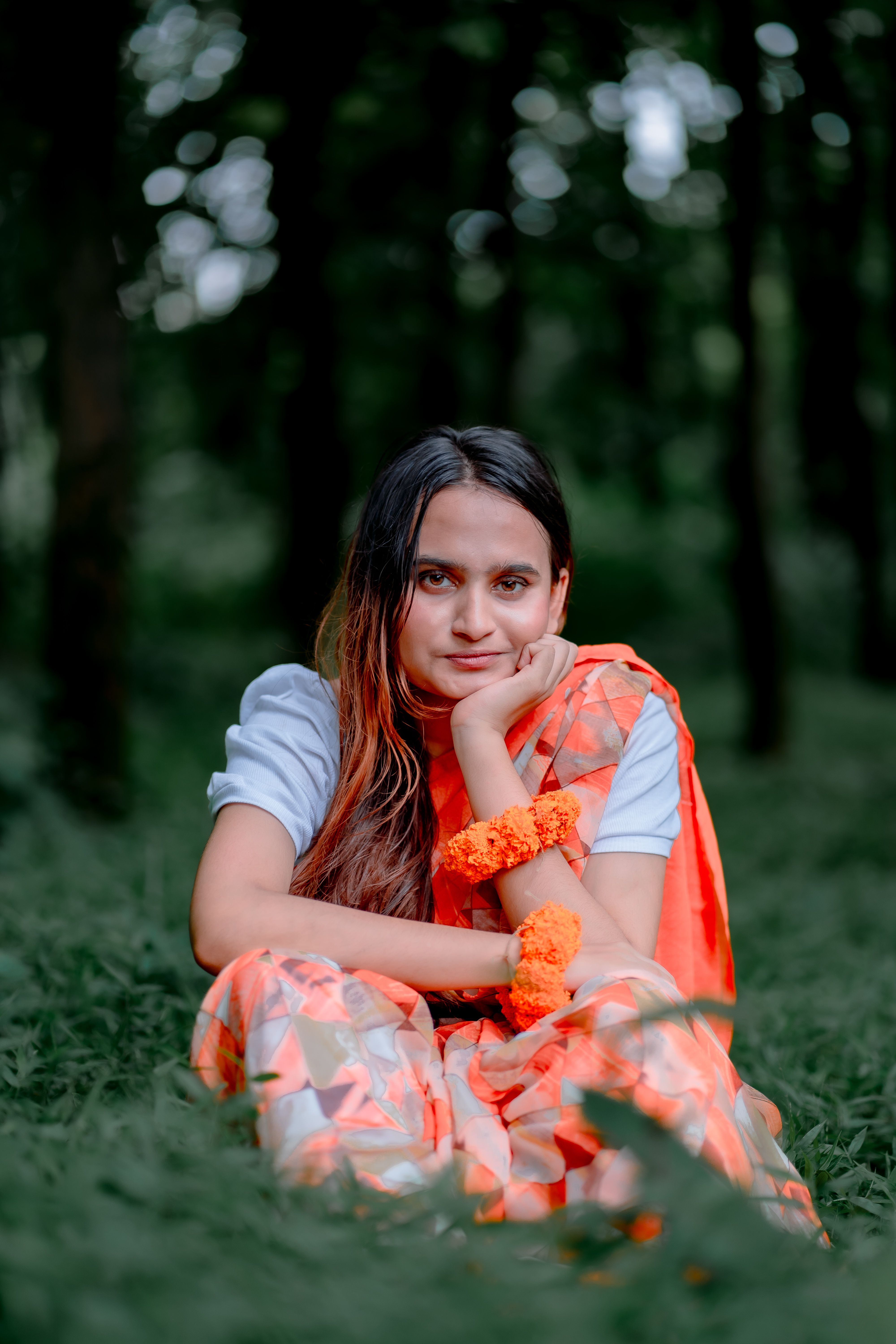 A woman wearing an orange sari sits peacefully in a forest, surrounded by greenery.