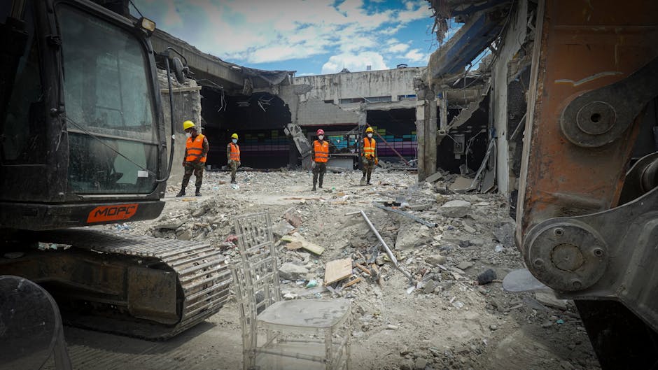 Construction workers assess a wrecked site with heavy machinery amidst rubble and debris.