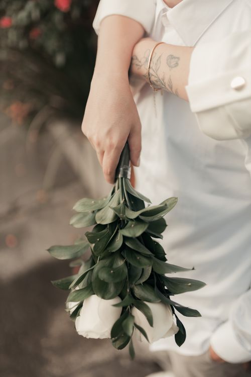 Free A hand holding a bouquet of white roses with greenery, featuring visible tattoos. Soft light and neutral tones. Stock Photo