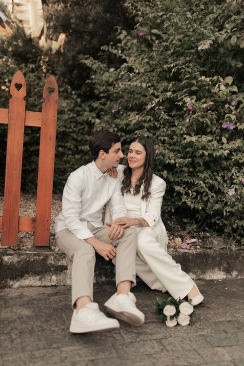 Free A happy couple in white outfits relaxes in a garden, surrounded by flowers and a wooden fence. Stock Photo