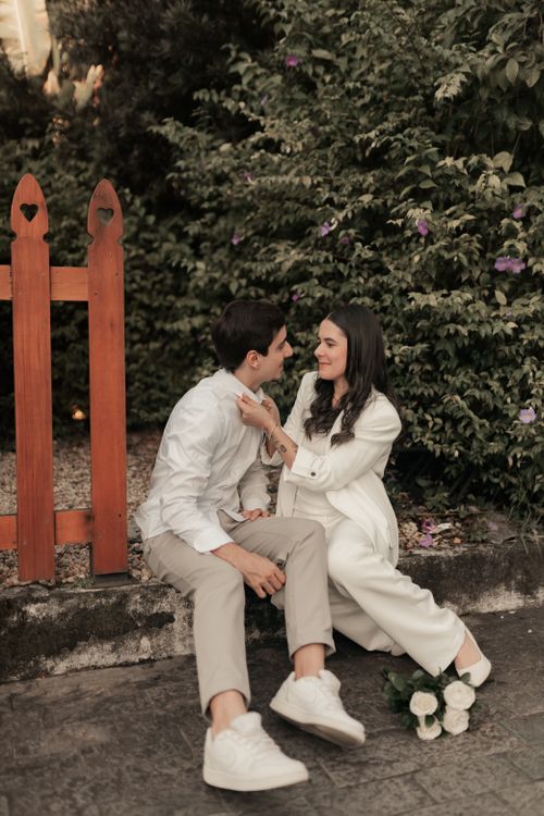Free A couple sitting by a red fence surrounded by lush greenery, sharing a tender moment outdoors. Stock Photo