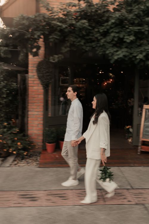 Free A young couple strolls holding hands past a leafy cafe on a sunny day. Stock Photo