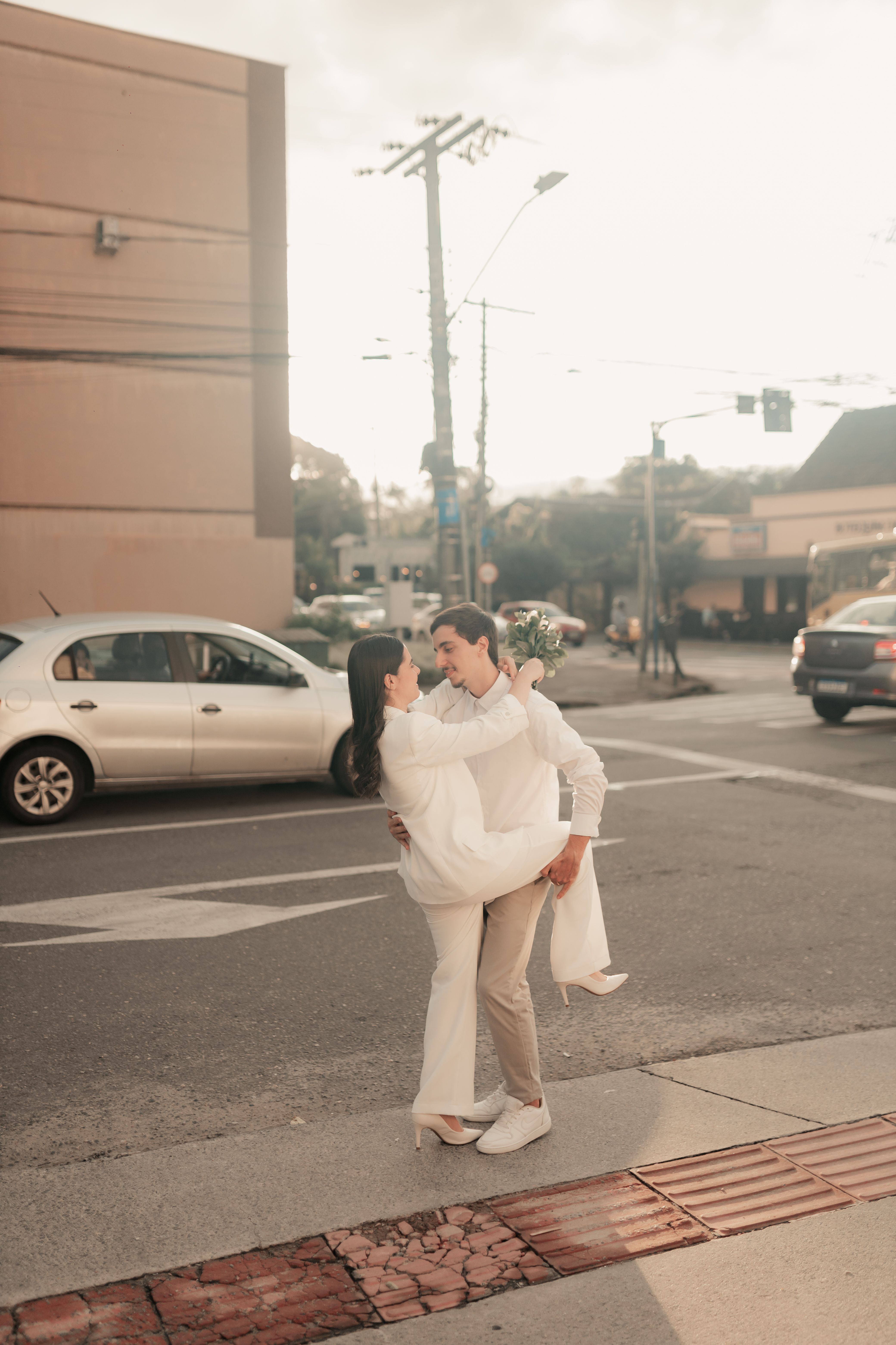 Happy couple in white attire embracing on a city street at sunset, creating a warm, joyful moment.