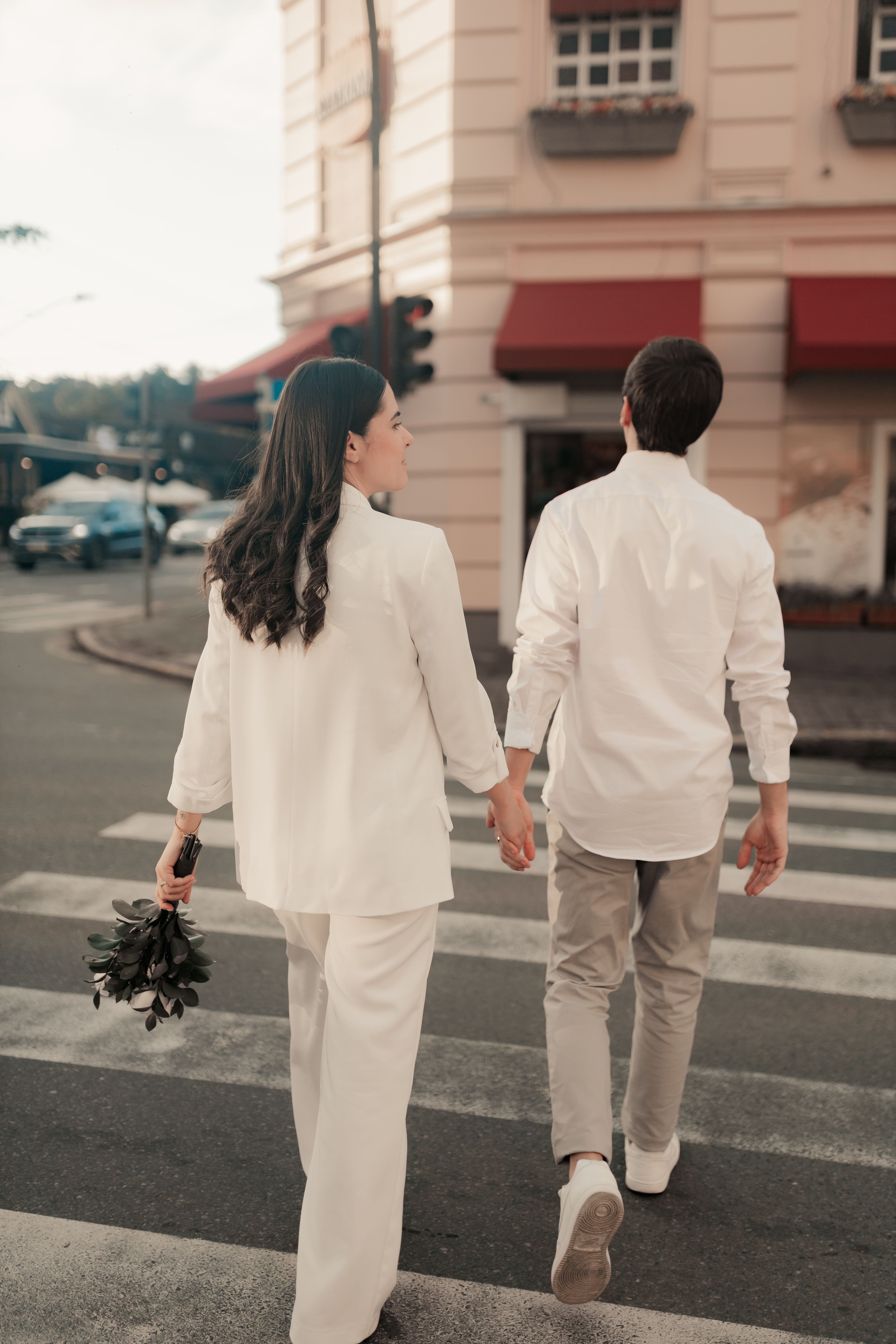A couple holding hands walking across a city crosswalk, wearing matching white outfits.