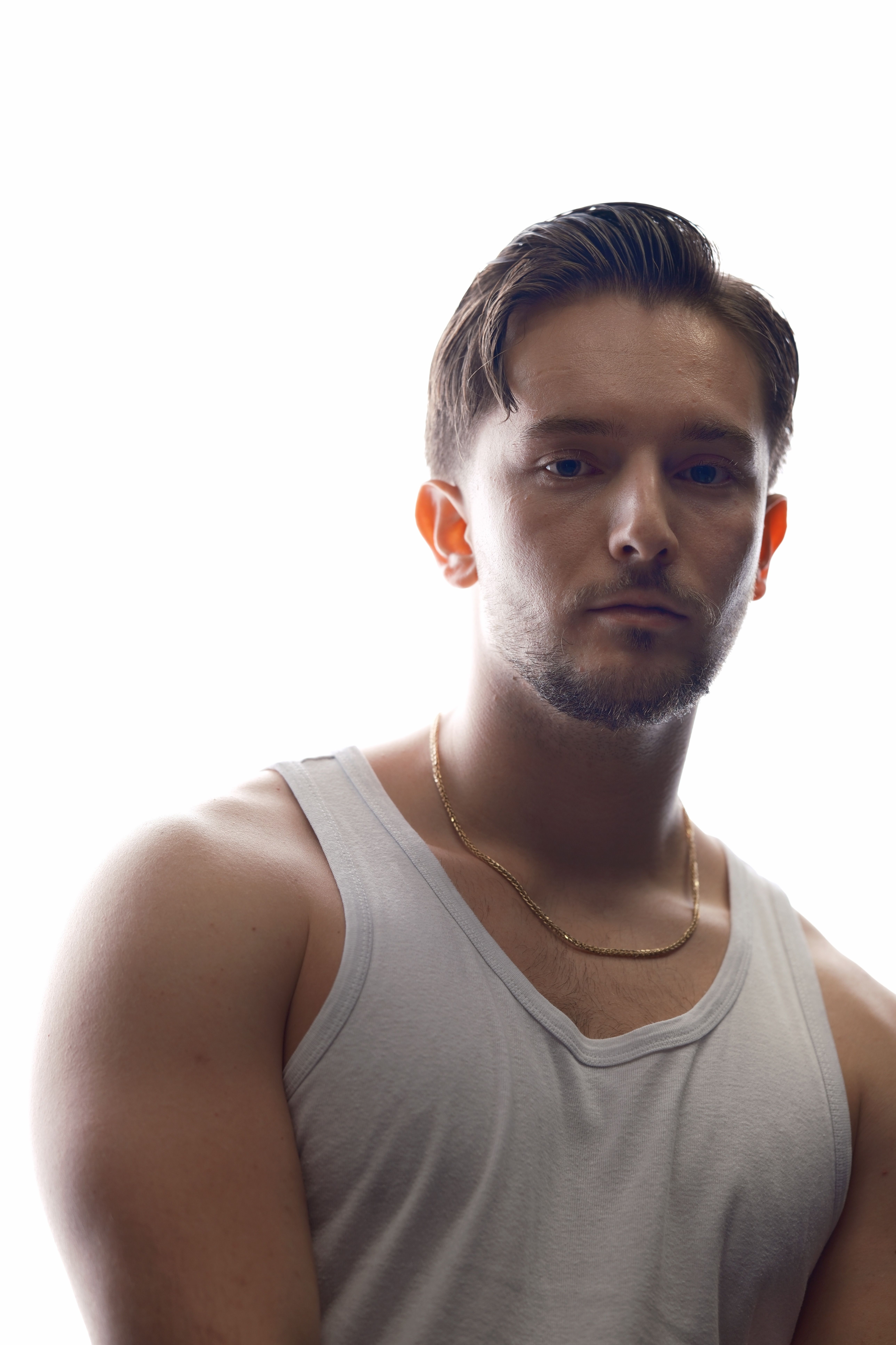 Dramatic portrait of a young man with short hair against a bright background, wearing a white tank top.