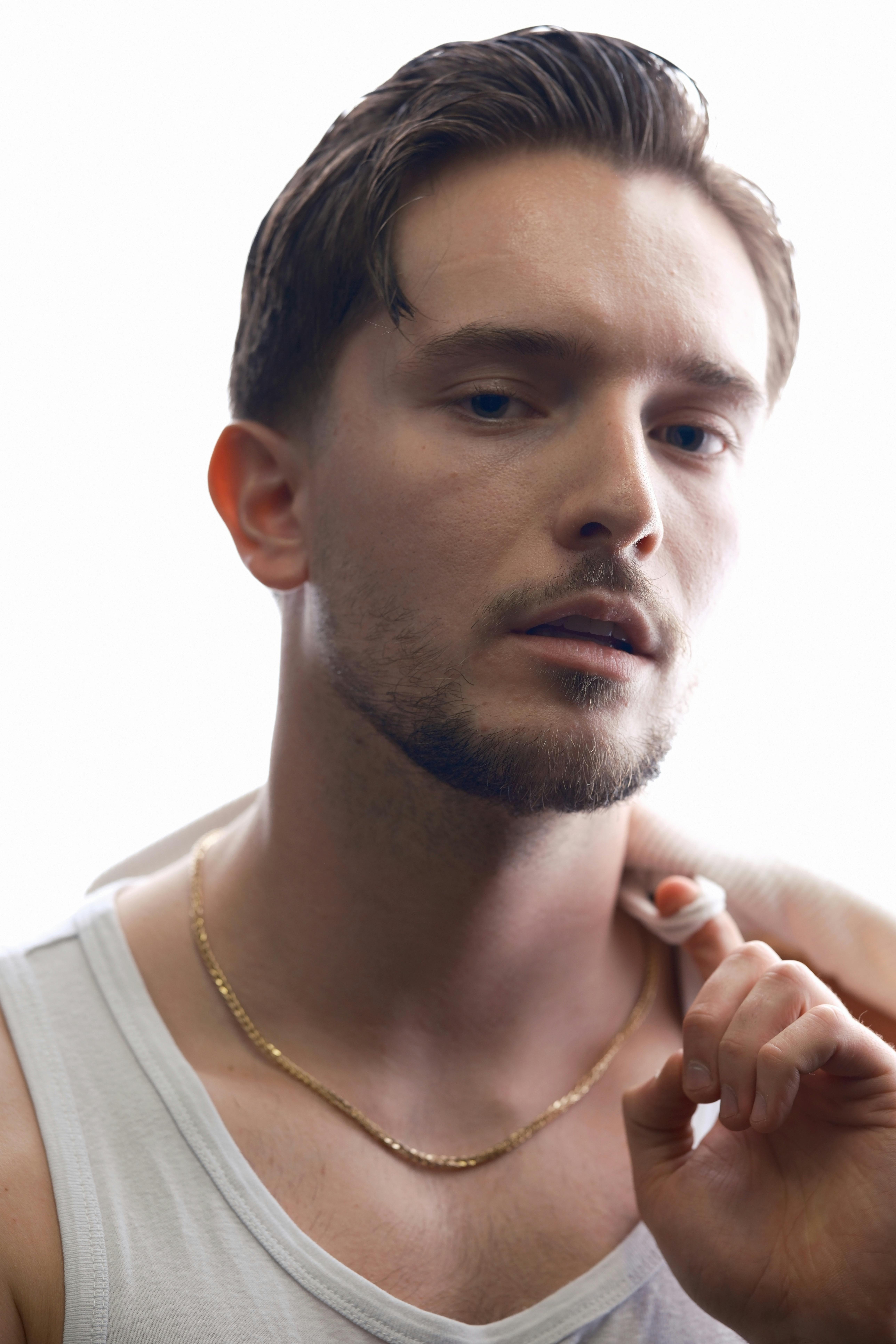 Portrait of a young Caucasian man wearing a white tank top and gold necklace, with a soft expression.