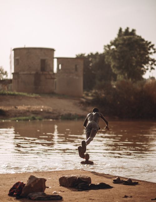 Free A man gracefully jumps into a river at sunset with rustic structures in the background. Stock Photo