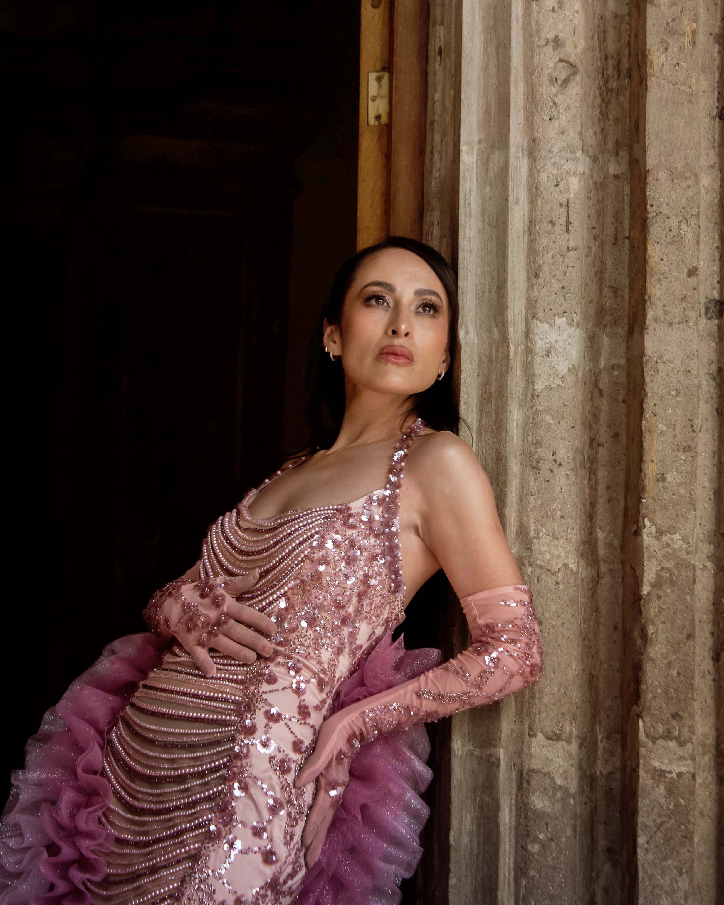 A sophisticated woman in a pink sequined gown poses gracefully against a stone wall, exuding elegance.