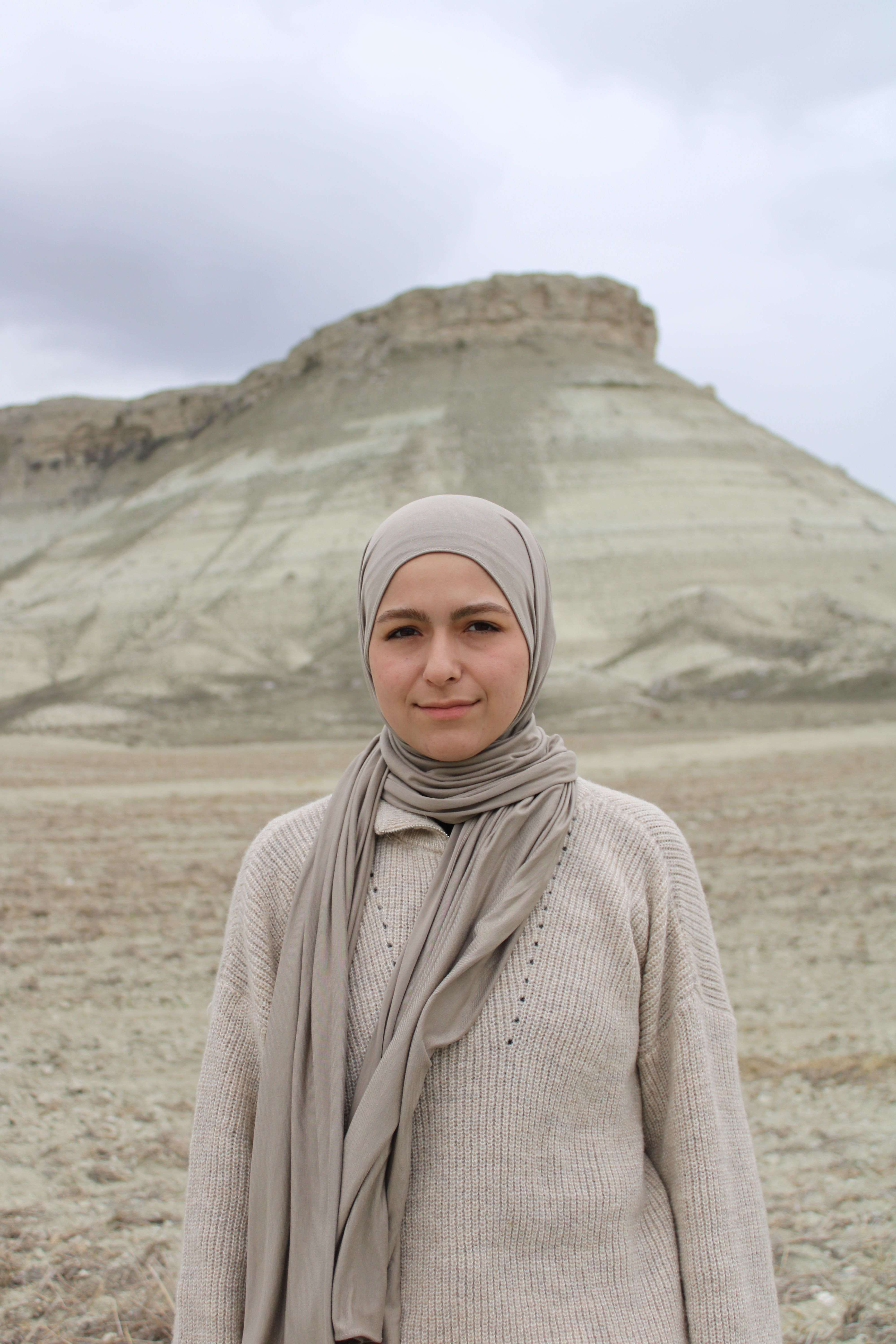 A woman in neutral attire stands in front of a mountainous landscape.