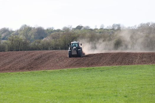 A tractor plows a field on a sunny spring day, creating dust in the air.