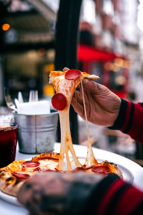 Free Close-up of a hand pulling a slice of pepperoni pizza with stretchy cheese outdoors. Stock Photo