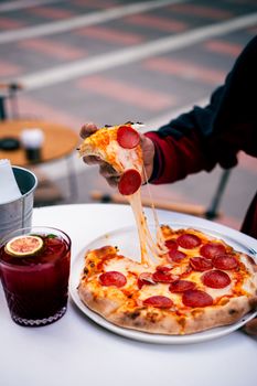 Close-up of pepperoni pizza with stretched cheese and a refreshing beverage outdoors.