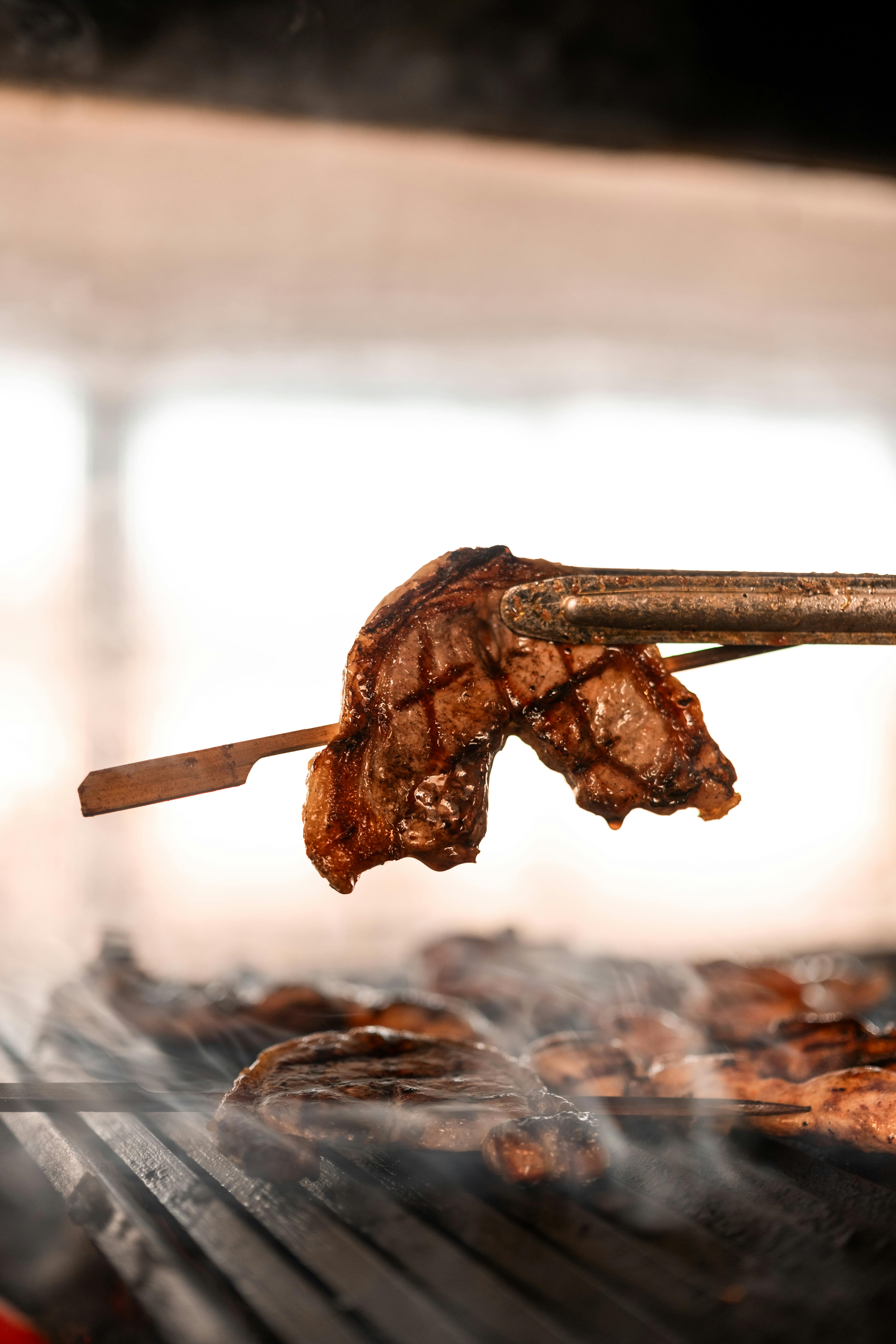 Close-up of a juicy grilled steak on barbecue tongs, cooking on a smoky grill.