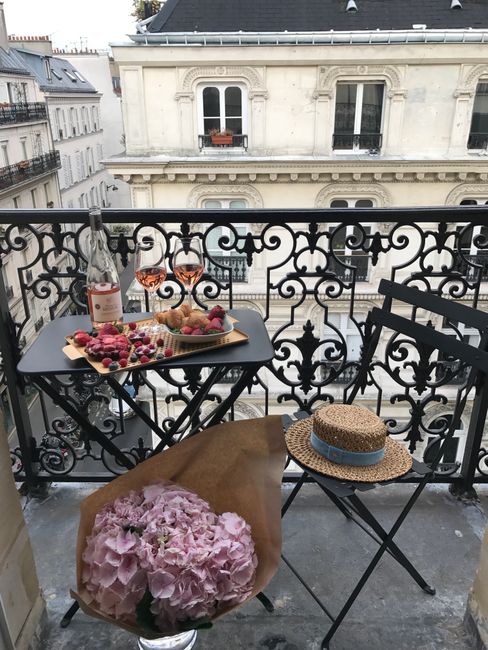 Cozy Parisian balcony setup with wine, snacks, and flowers on a sunny day.