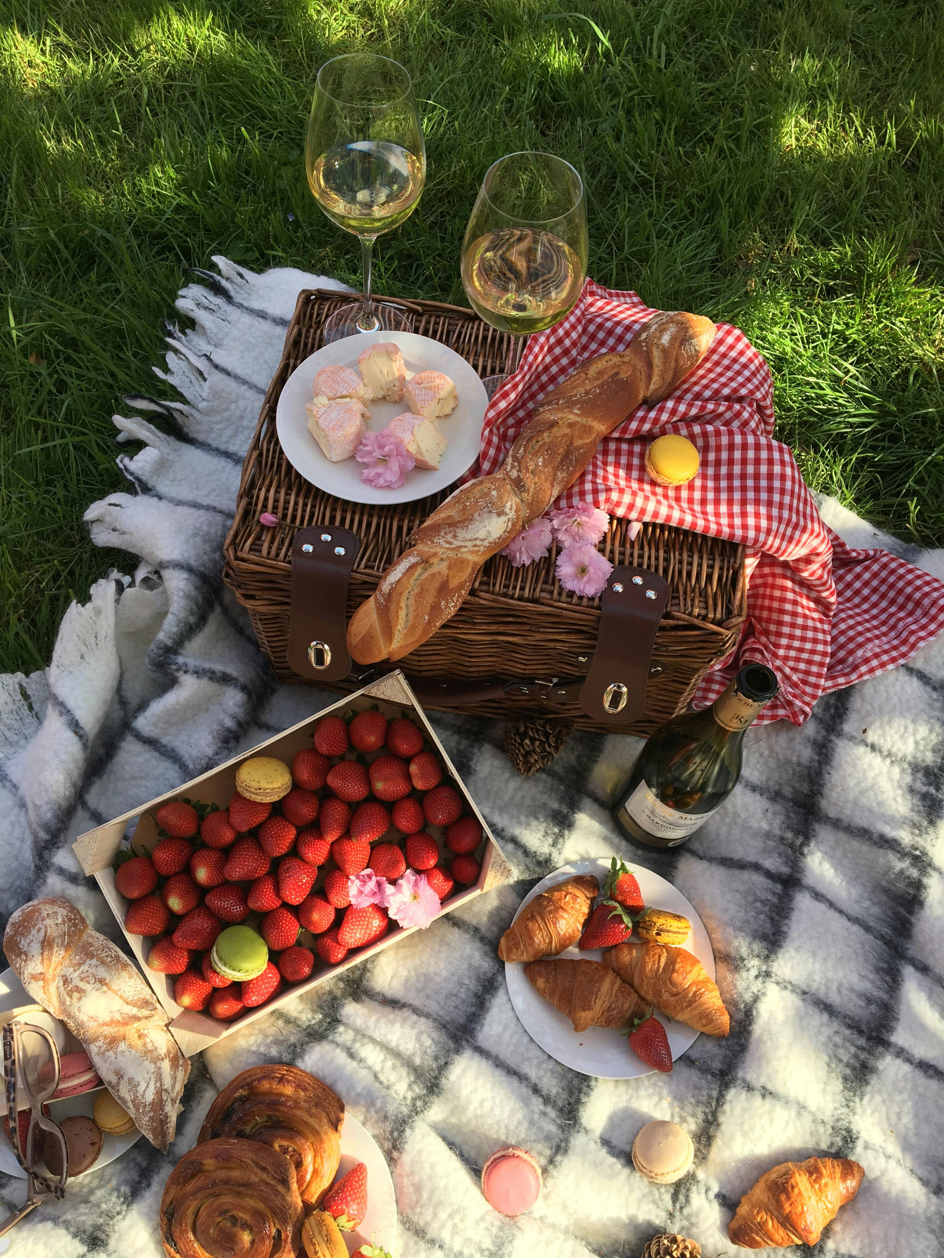 A summer picnic setup featuring baguettes, strawberries, croissants, wine, and macarons on a blanket.