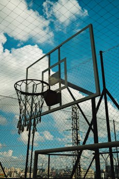 Outdoor basketball court with hoop against a cloudy blue sky, featuring an urban setting and sports theme.