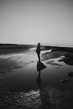 A moody black and white photo showing a solitary figure at the beach during twilight.