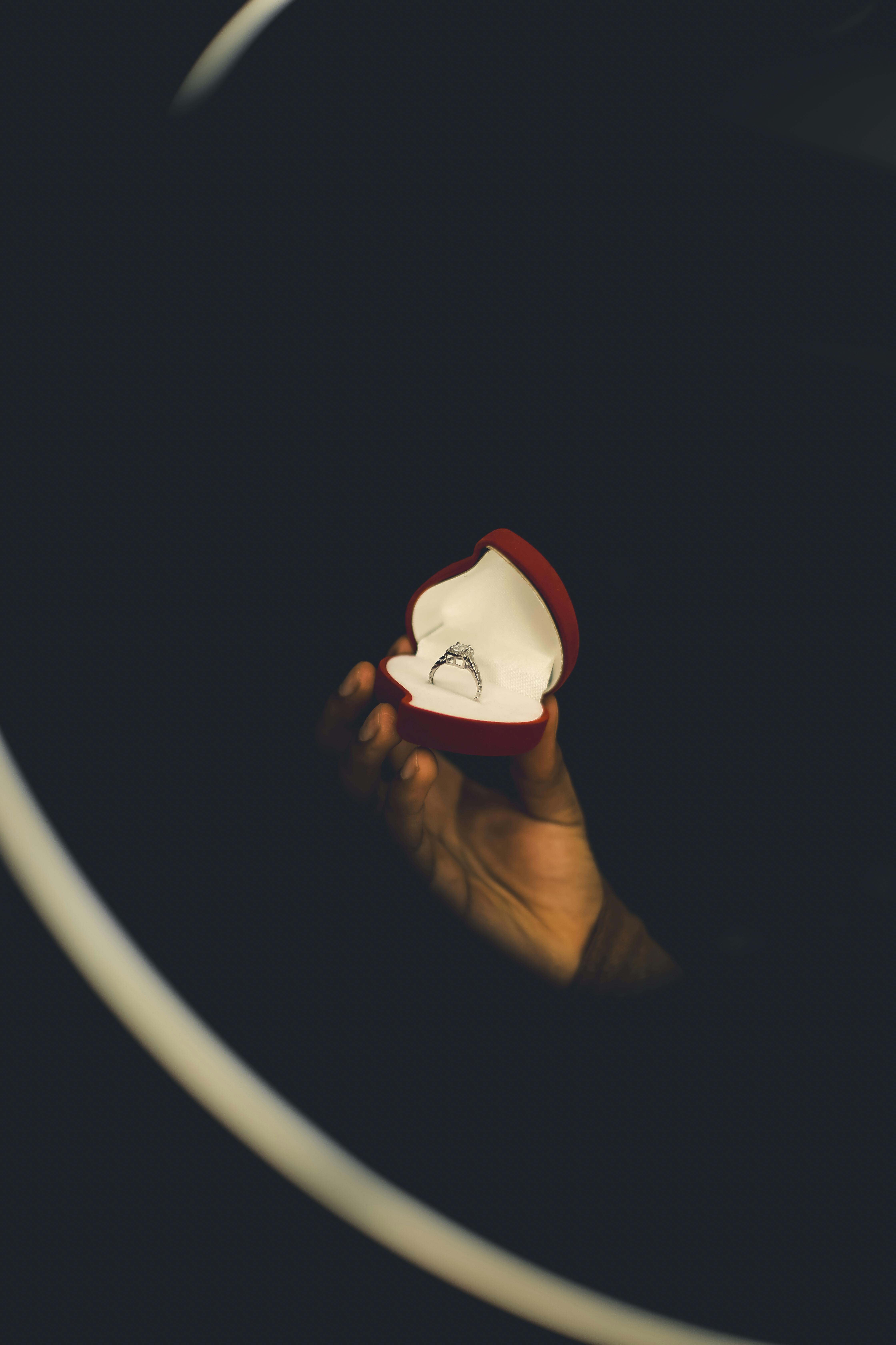 Gratis Sebuah tangan memegang kotak cincin pertunangan merah terbuka dalam pencahayaan dramatis. Foto Stok