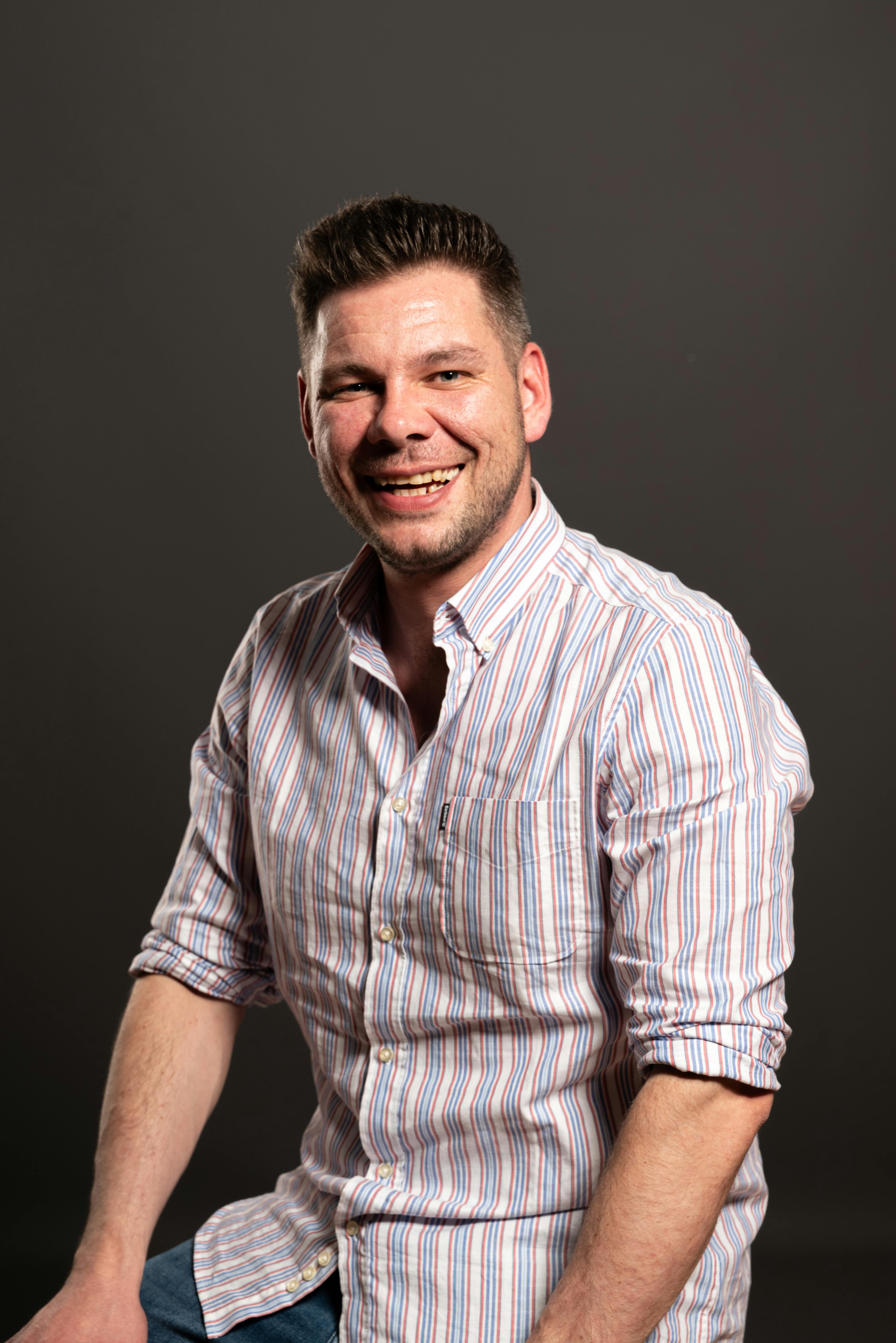 Portrait of a smiling adult man in casual striped shirt with dark backdrop, exuding warmth and friendliness.