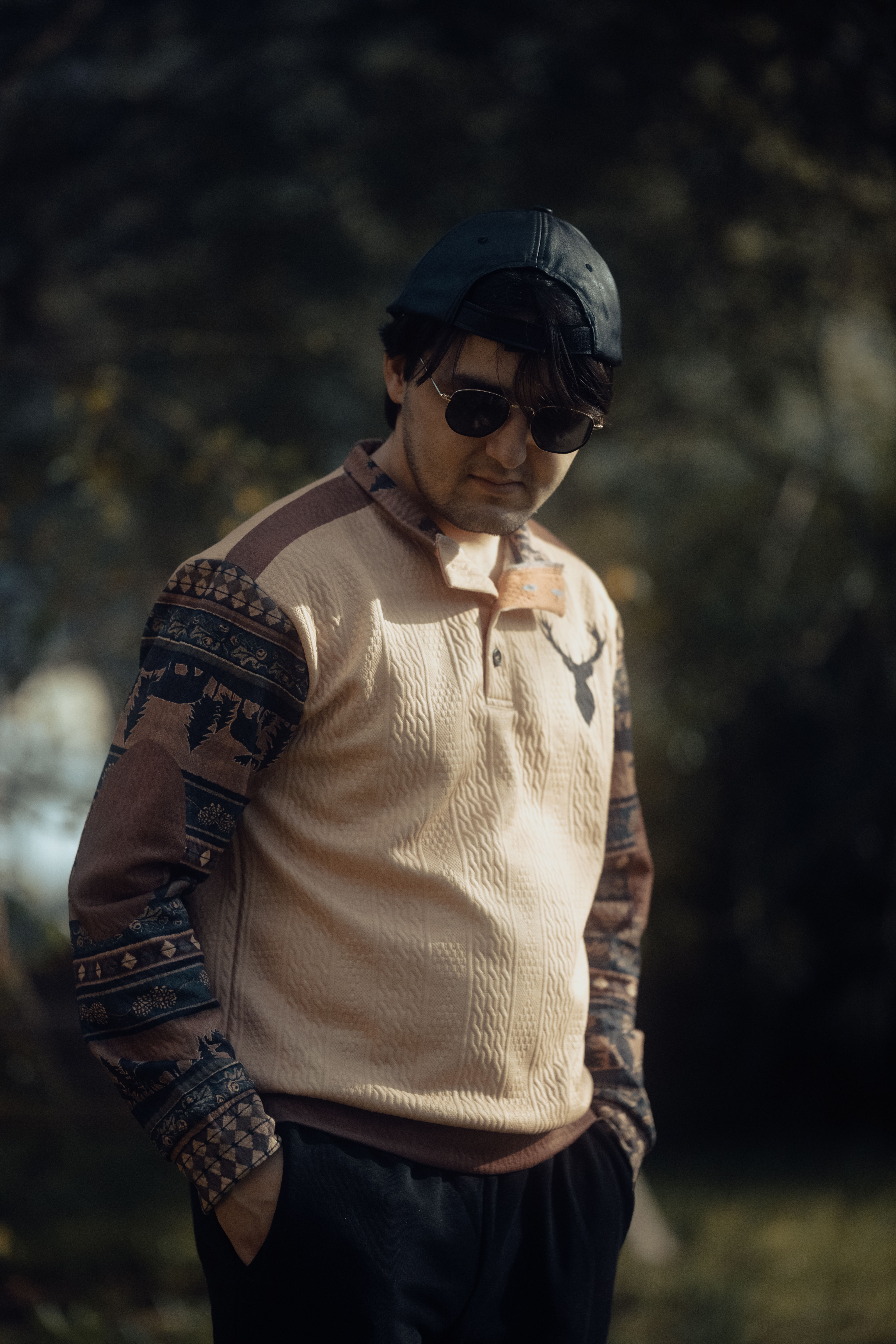 Man wearing a stylish sweater with patterns and sunglasses standing outdoors in a moody setting.