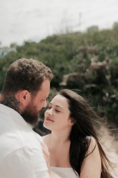 A couple shares a tender moment outdoors on a sunny beach with lush greenery.