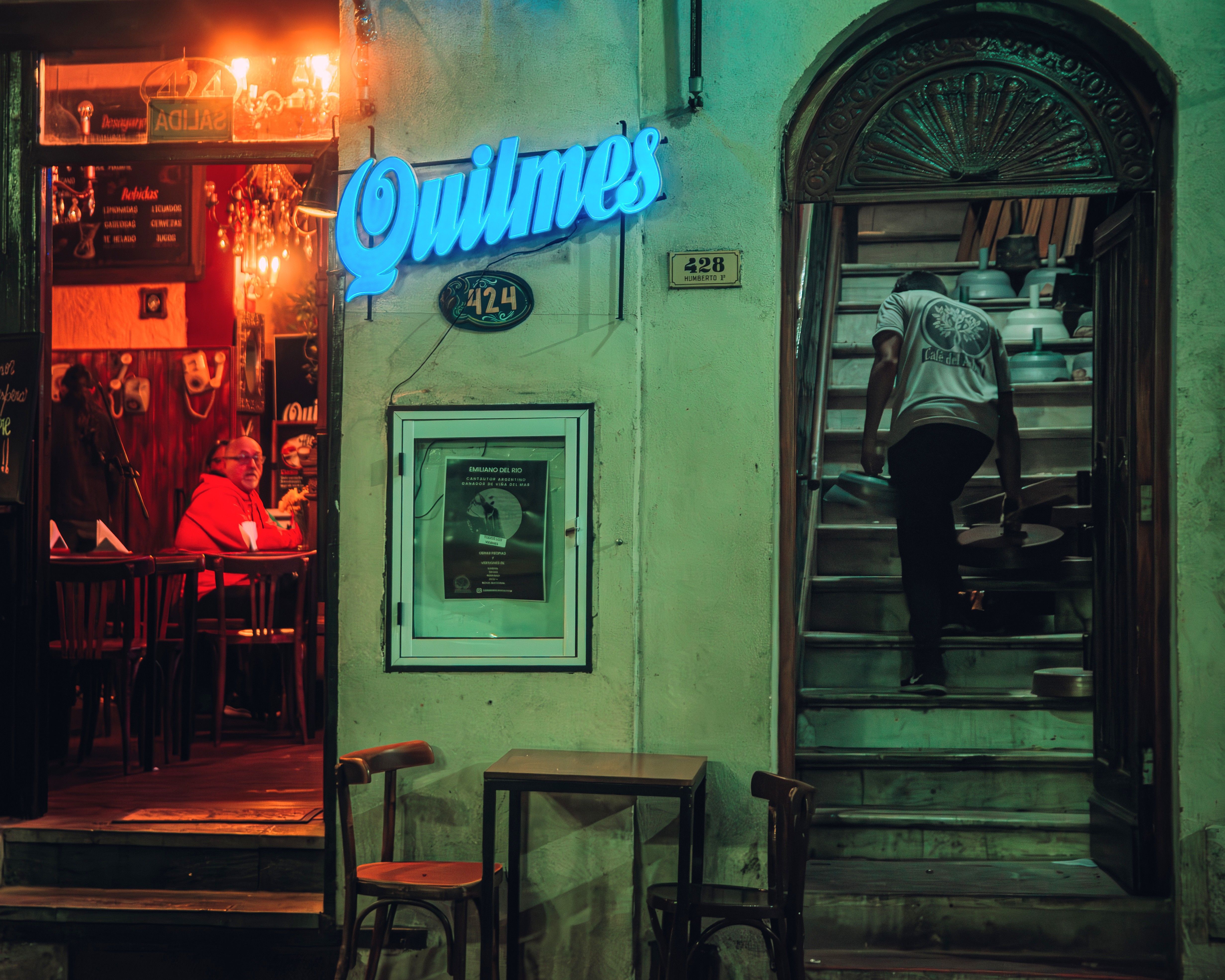 Charming bar entrance with neon sign, warm lighting, and a man climbing stairs at night.