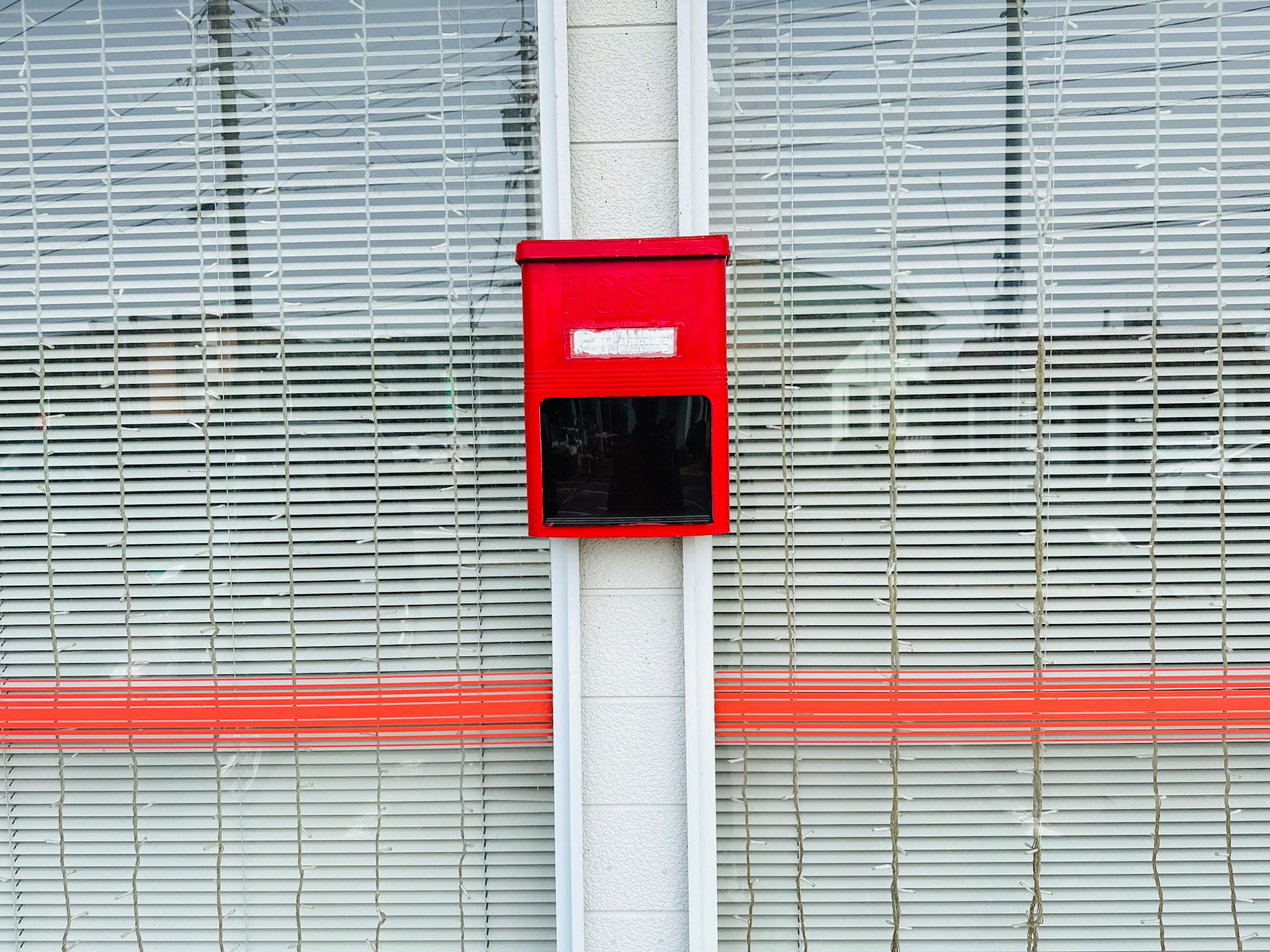 Red post box on a street wall with striped window blinds behind. Minimalist urban style.