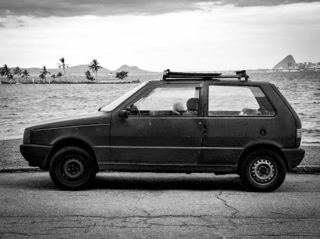 Black and white photo of a retro car by the ocean with mountain backdrop.