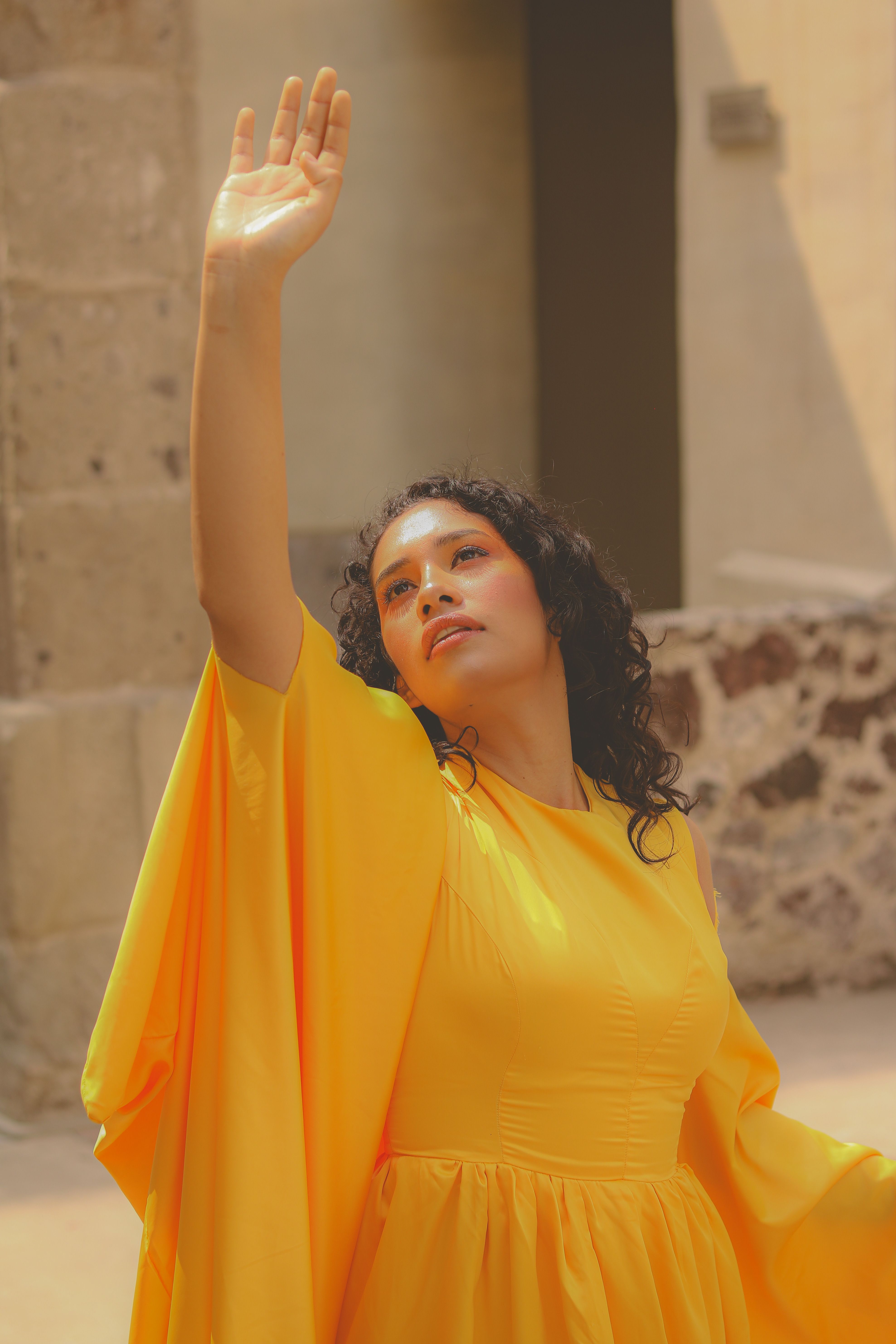 Woman in a yellow dress reaching upwards, posing outdoors with a sunny backdrop.