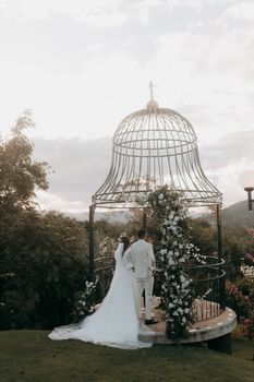 Bride and groom in elegant attire at a floral-decorated gazebo, outdoors.