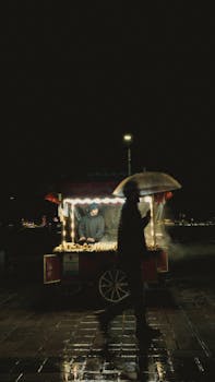Night scene with a street vendor and a passerby holding an umbrella, creating a moody ambiance.