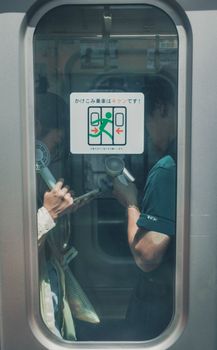 Two individuals on a Tokyo train using handheld devices, seen through a window, reflecting urban life in Japan.