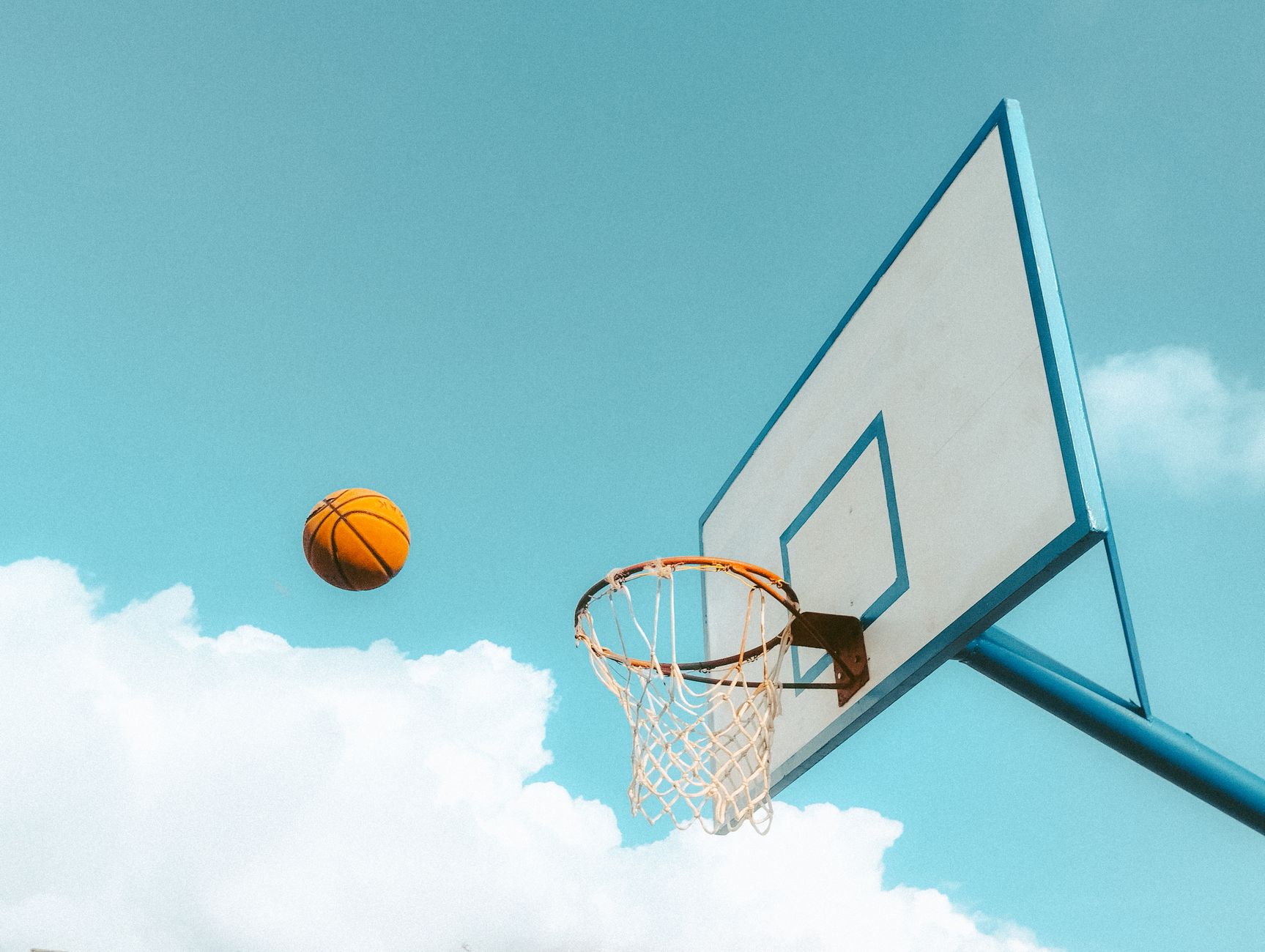 A basketball is about to make a swoosh into the net under a bright blue sky, symbolizing outdoor sports energy.